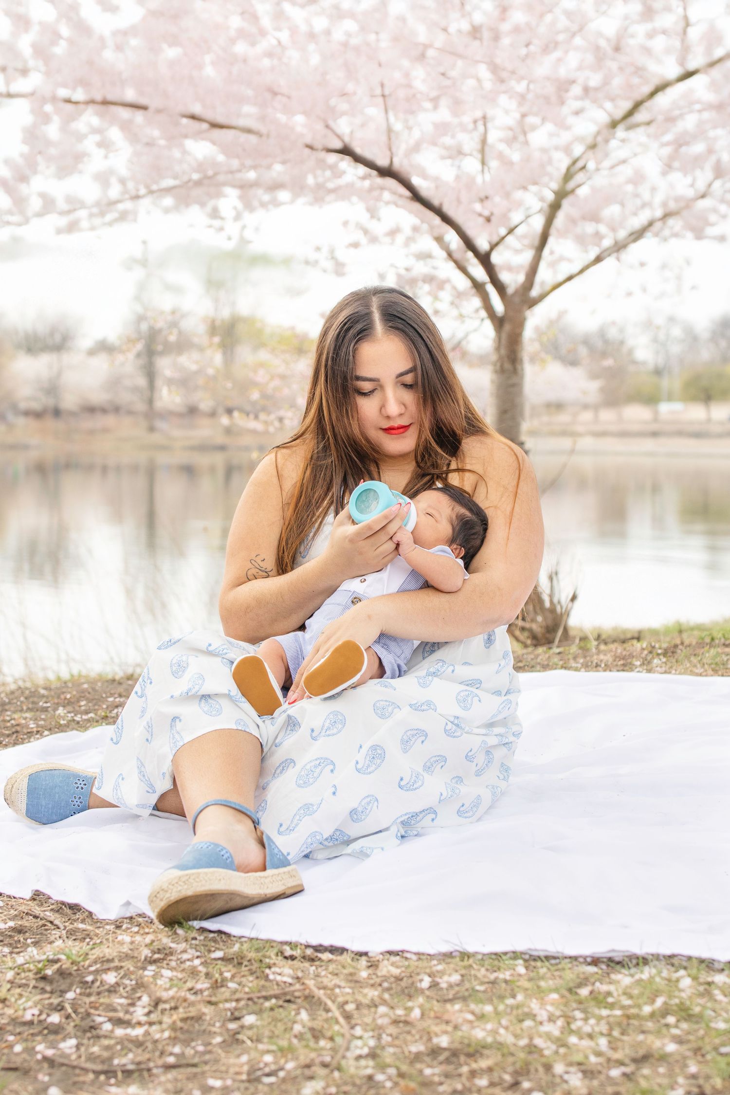 A tender moment on a picnic blanket under blooming cherry blossoms by a tranquil lake while feeding a baby.