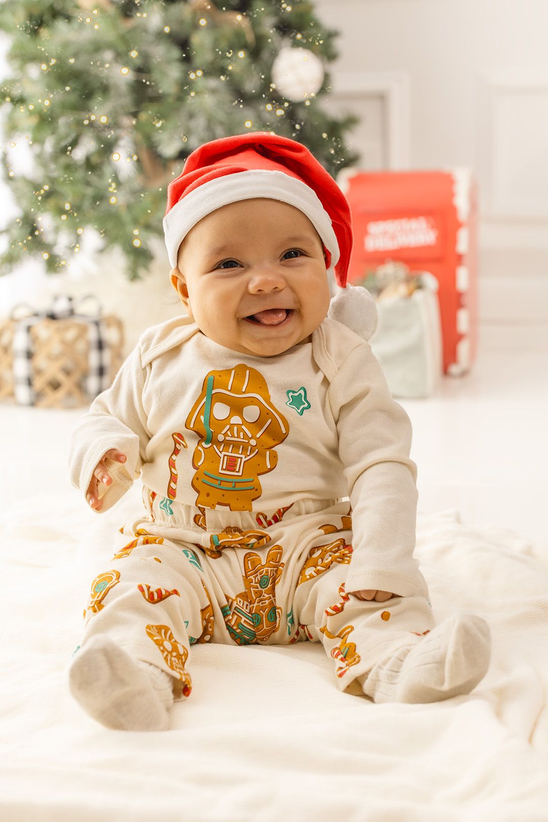 Baby wearing Santa hat and festive gingerbread outfit smiles during Christmas photo session with white decor.