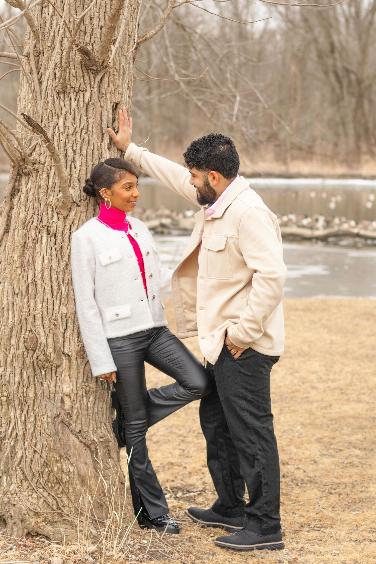 A couple poses casually against a tree trunk in coordinated winter outfits.