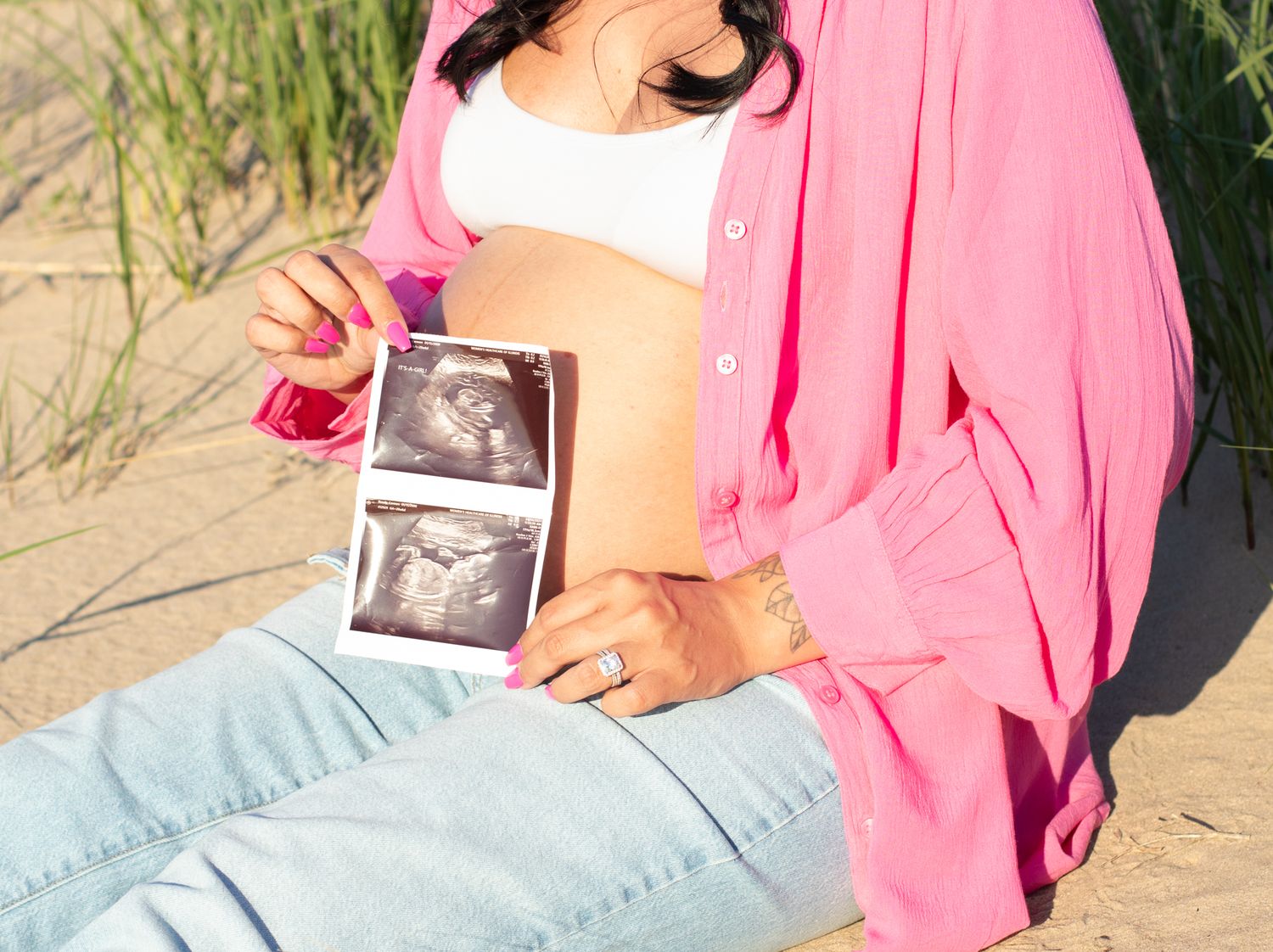 Expectant parent in pink cardigan holds ultrasound photos while sitting on beach dunes.