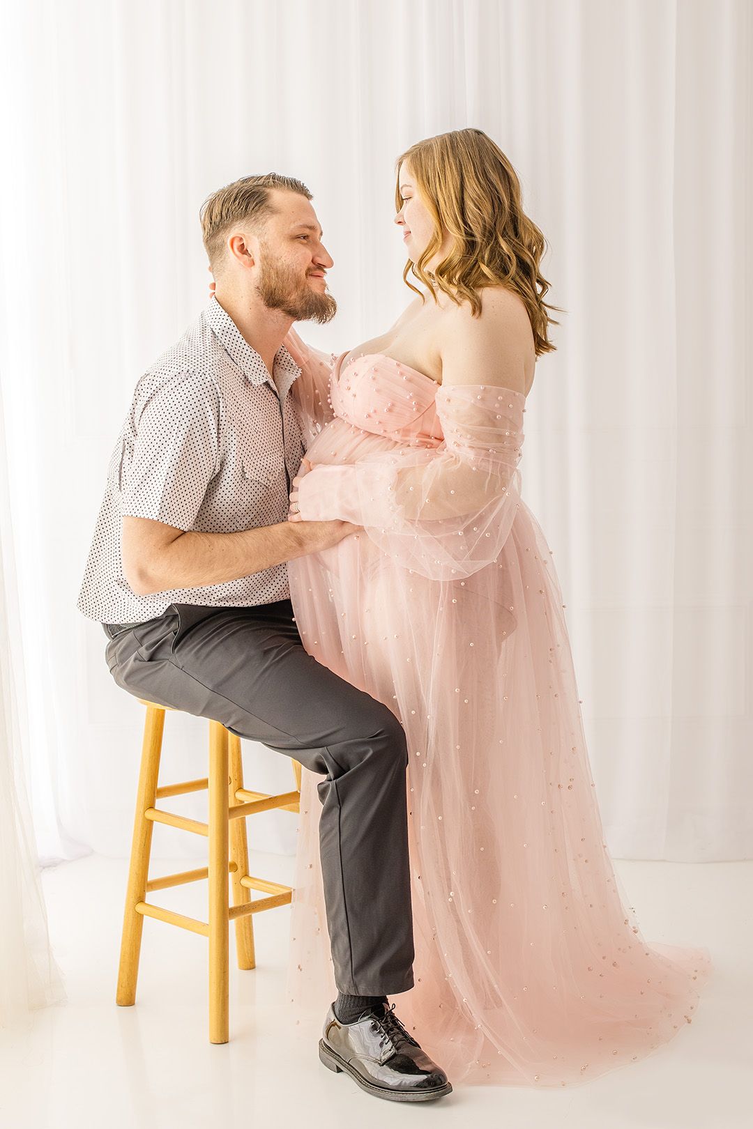 Romantic maternity photo featuring couple in pink tulle gown and casual attire against white backdrop with wooden stool.