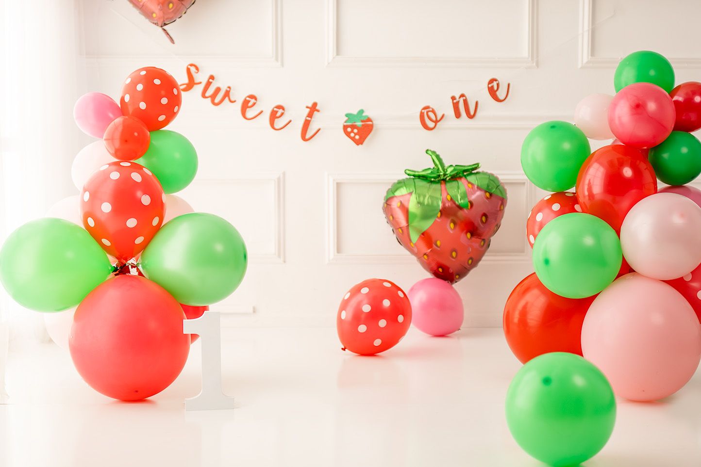 Strawberry first birthday party decor with red, green, and pink balloons and a sweet one banner.