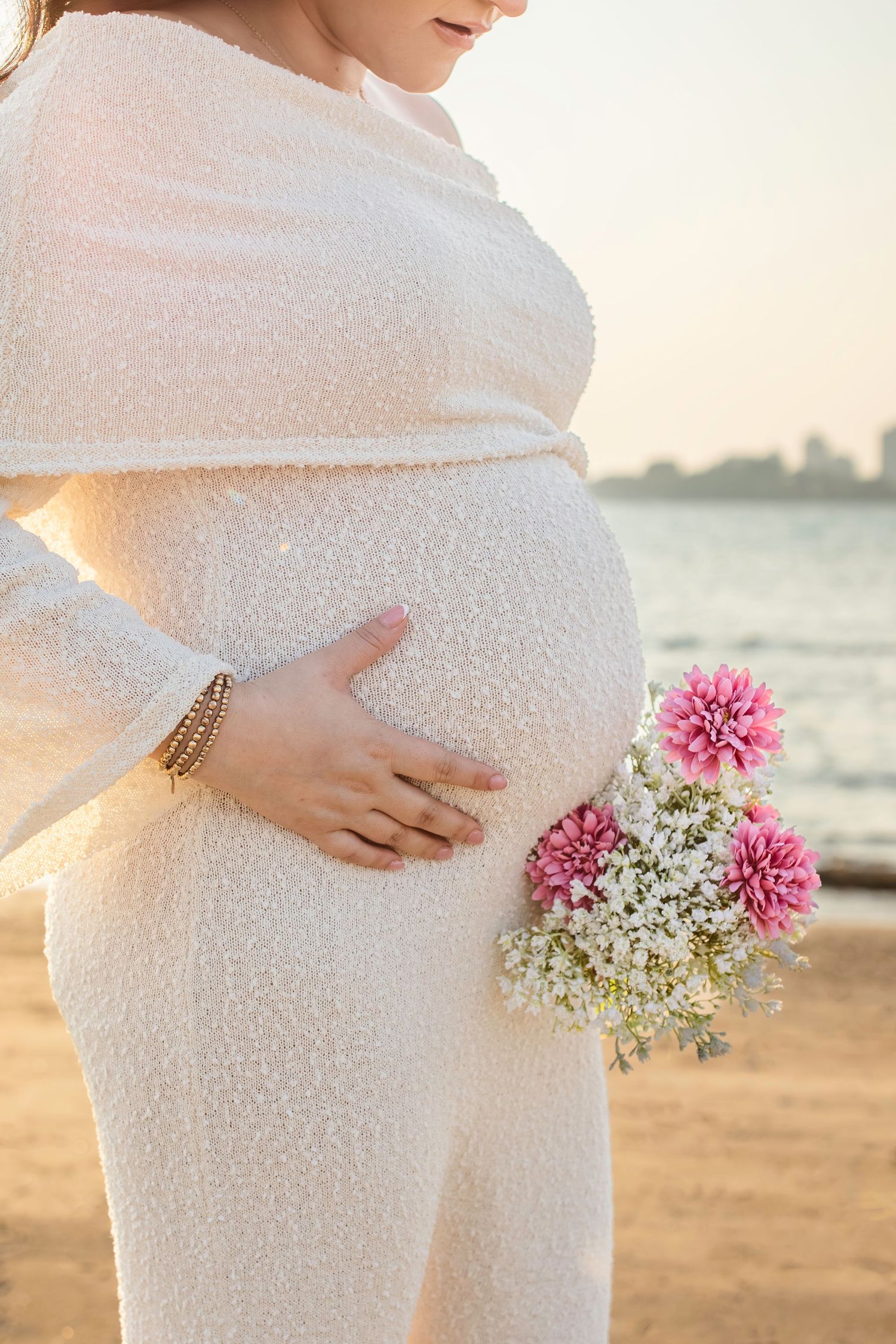 Maternity photos on beach showing baby bump with delicate pink and white flowers at sunset.