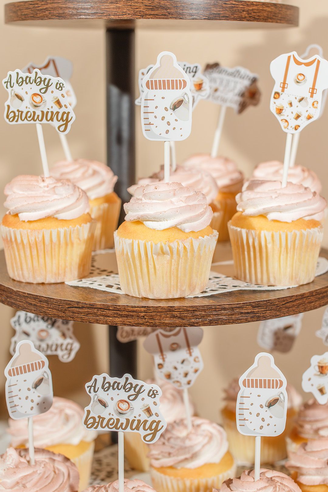 Baby shower cupcakes with pink frosting and 'A Baby is Brewing' toppers displayed on a wooden tiered stand.