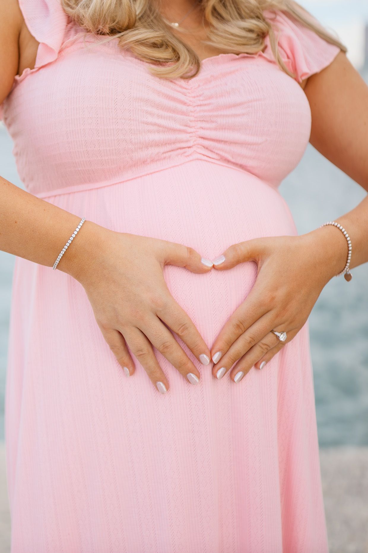 Pregnant woman in pink dress forming heart shape with hands on baby bump.