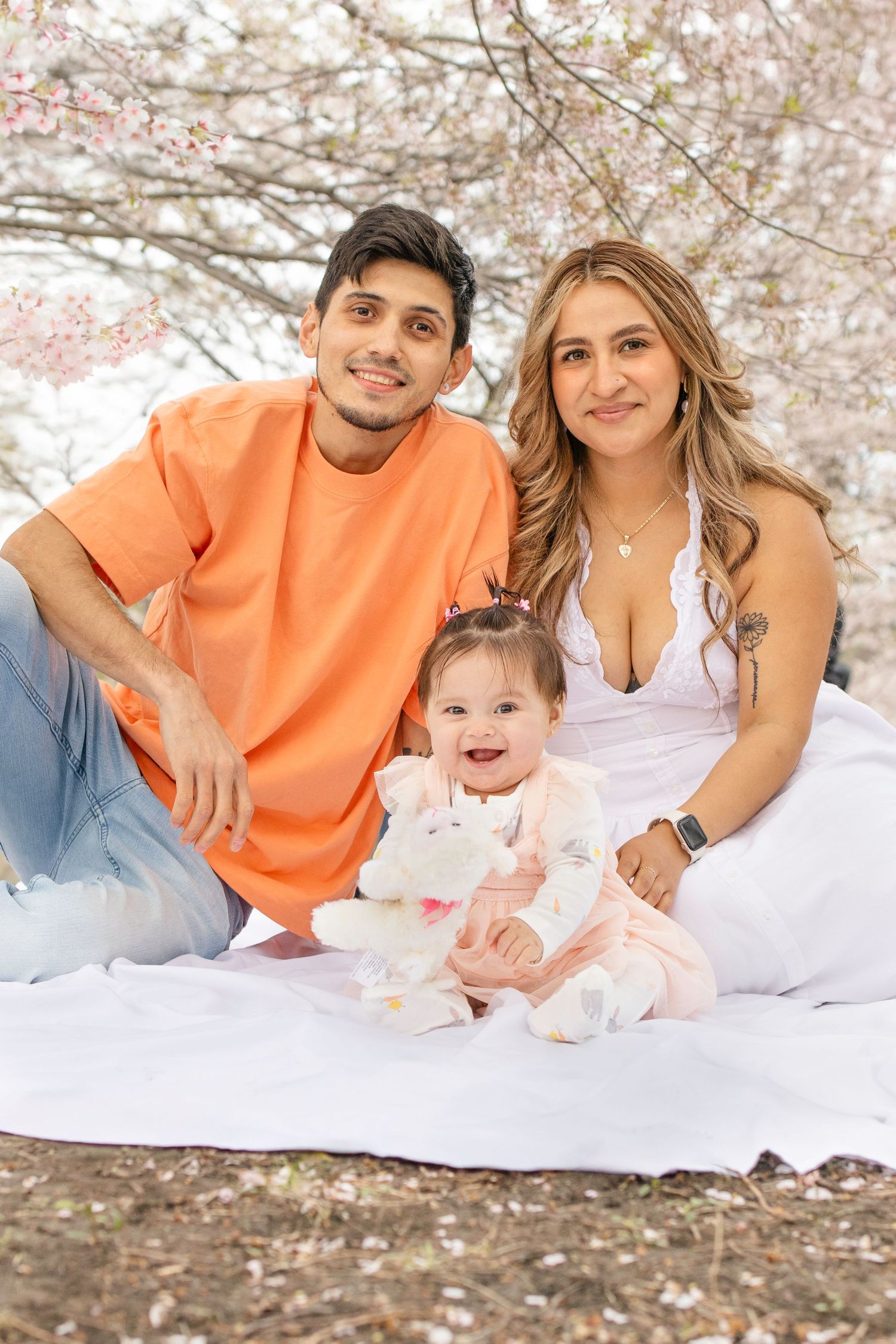 Young parents in orange and white outfits sitting with a happy baby on a blanket under blooming cherry blossom trees.