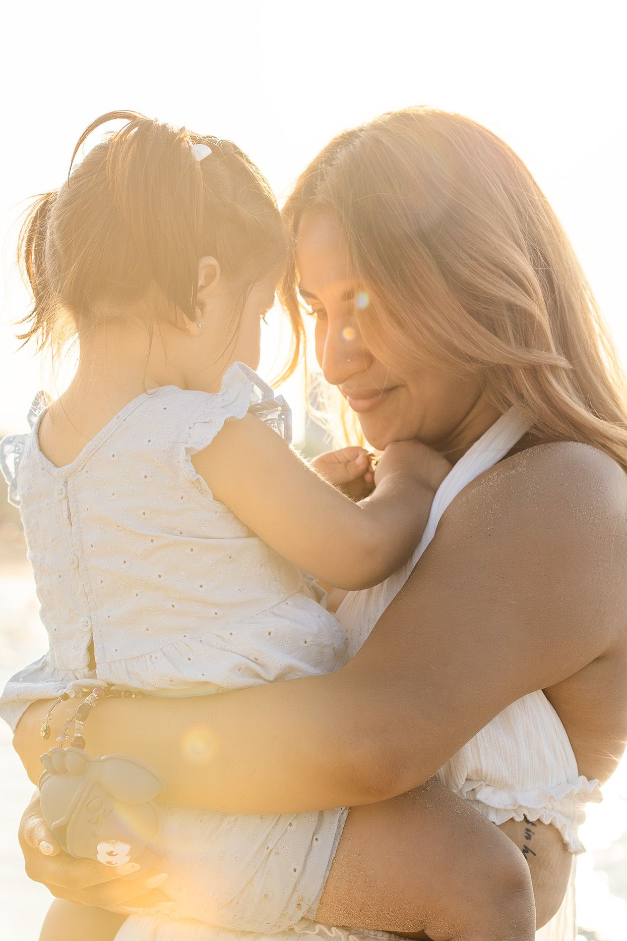 Tender moment of mother holding baby in white dress with warm golden sunlight glowing.