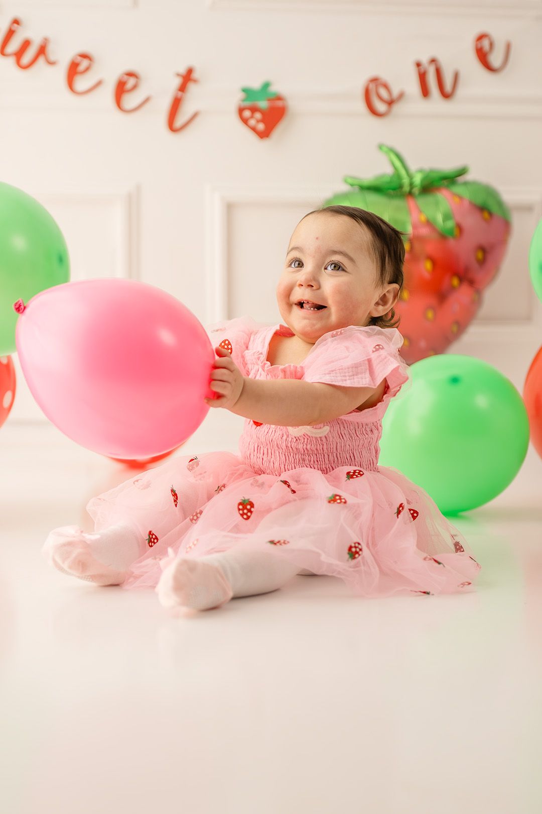 Baby girl in pink strawberry dress smiles at first birthday party with colorful balloons and sweet one banner.