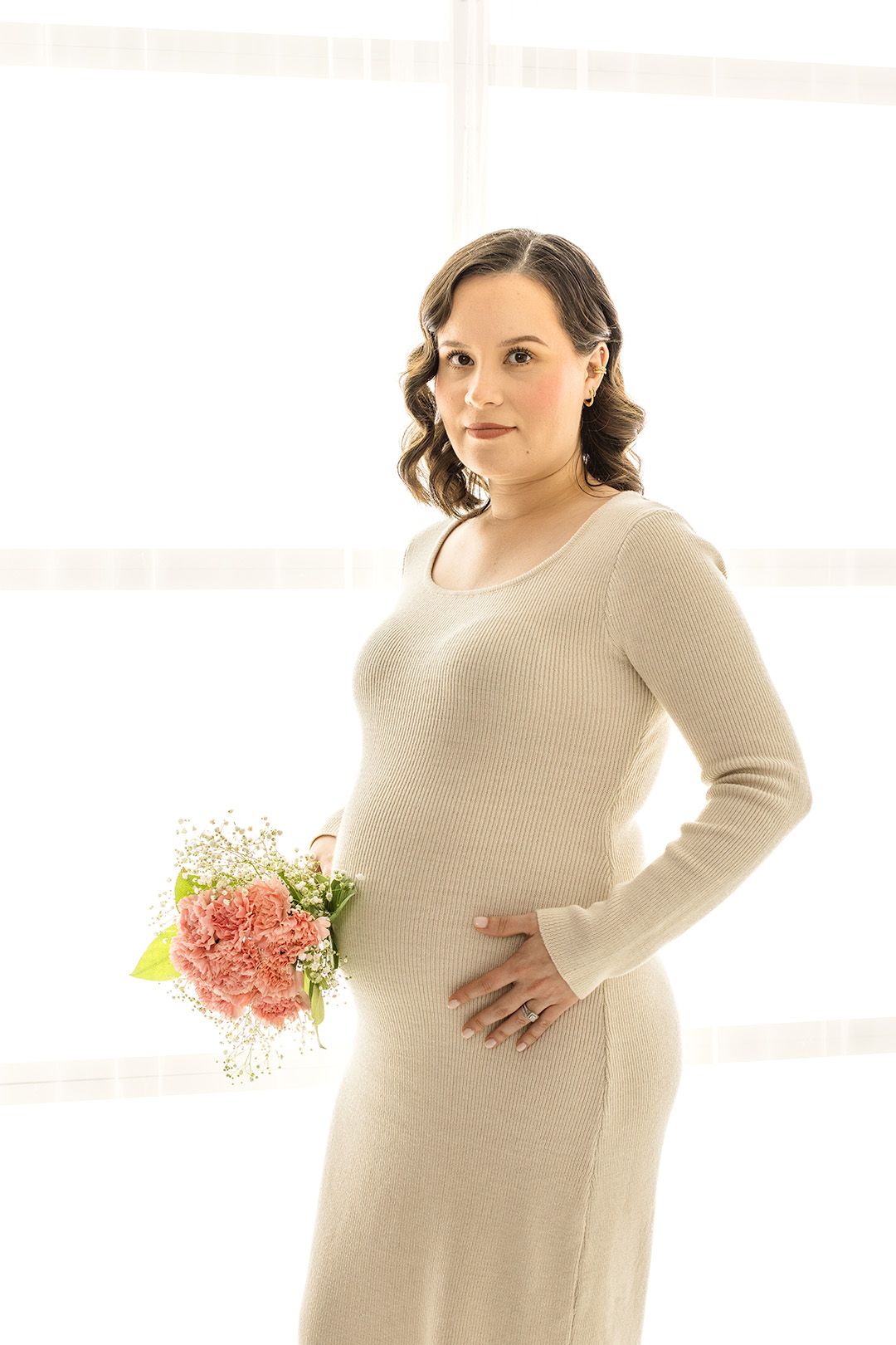 A pregnant woman in a beige knit dress holds pink flowers and cradles her baby bump in a bright, airy studio.
