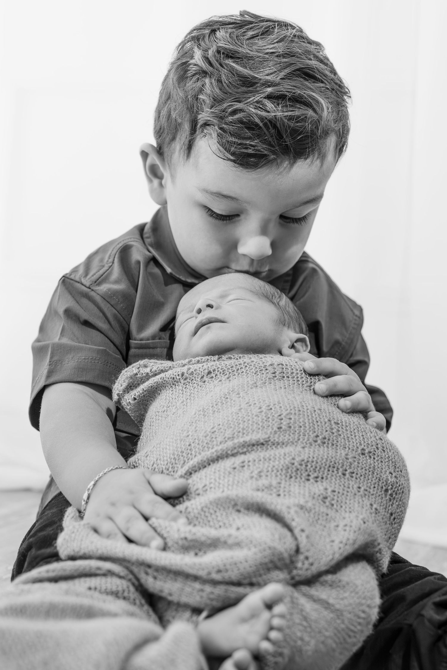 Black and white photo of a young child gently kissing a swaddled newborn baby.