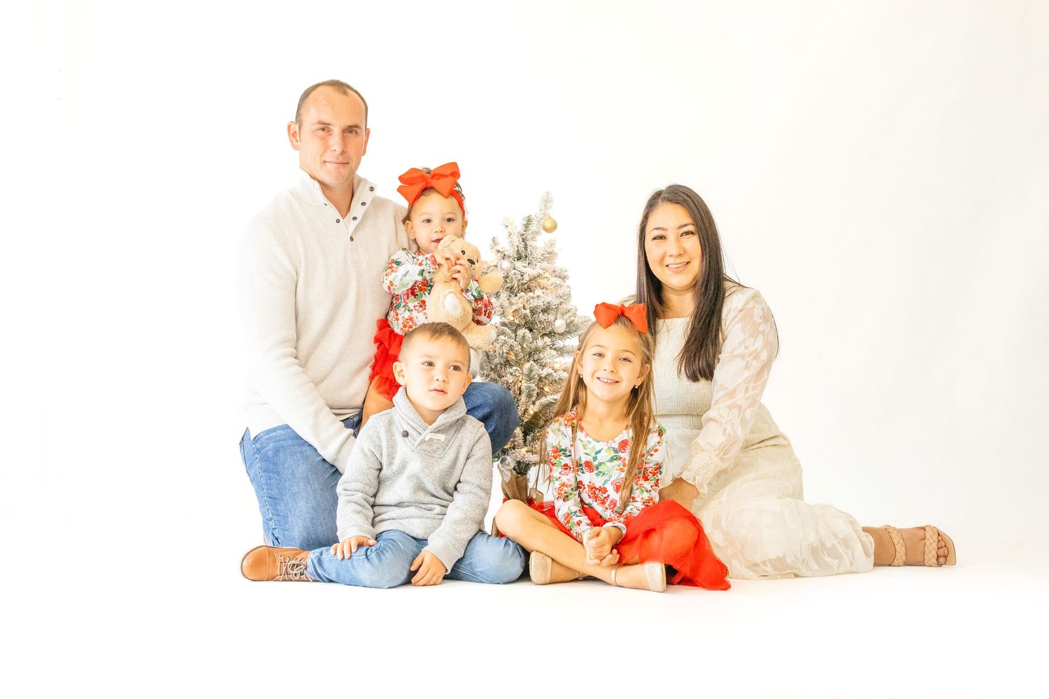 Family in white outfits poses together for holiday portrait with silver Christmas tree in studio setting.