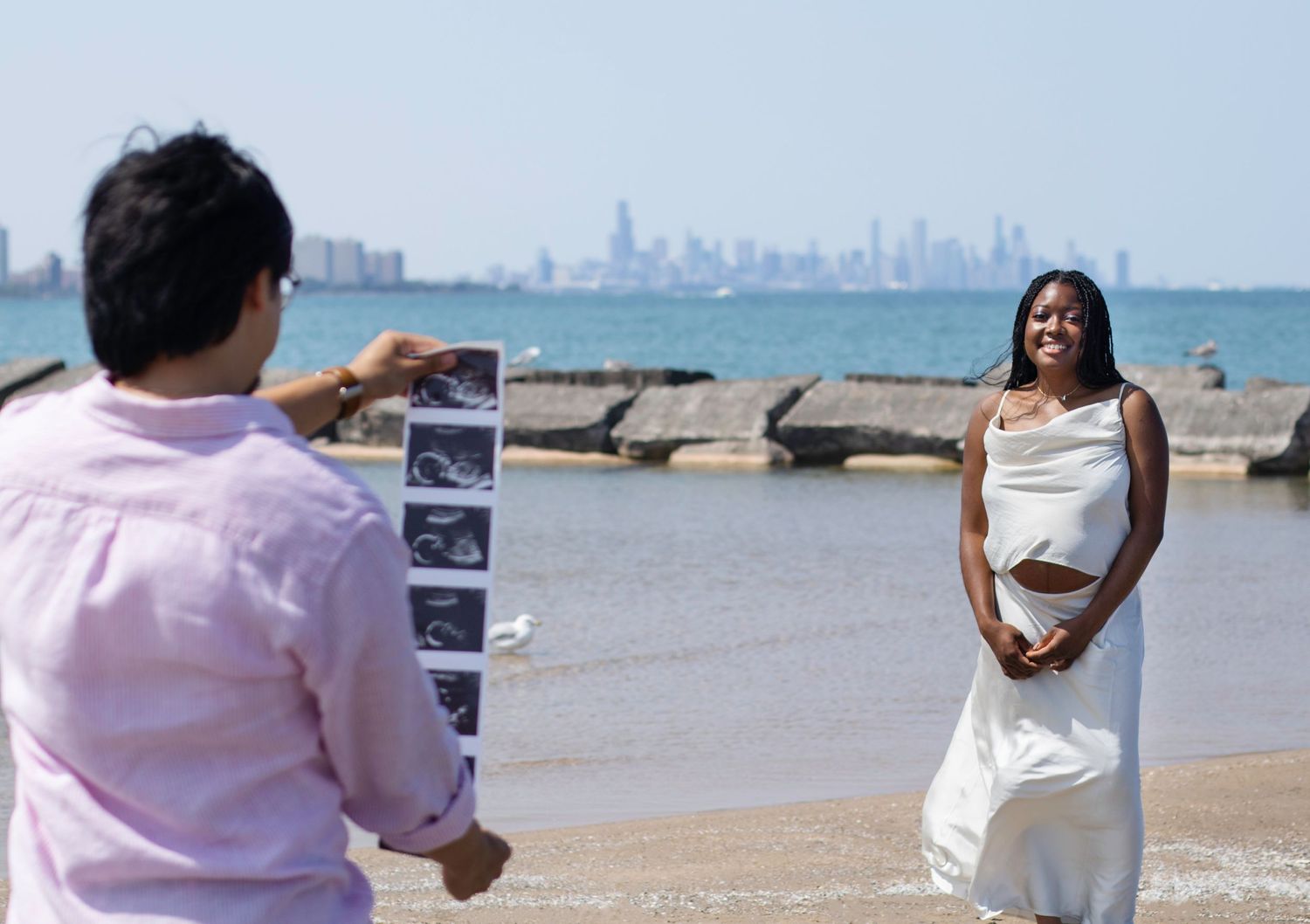 Photographer shows ultrasound photos during beachside maternity shoot with city skyline visible.