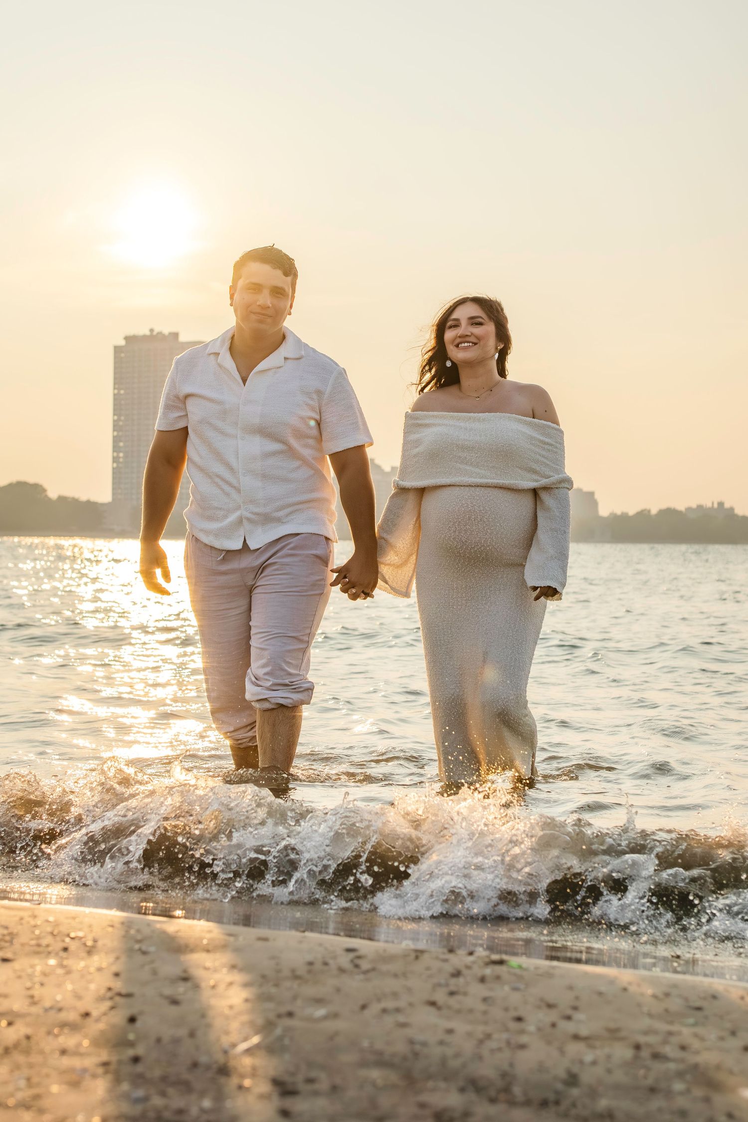 Couple walking along sunny beach shore with splashing waves and silhouette of city skyline during golden hour sunset.