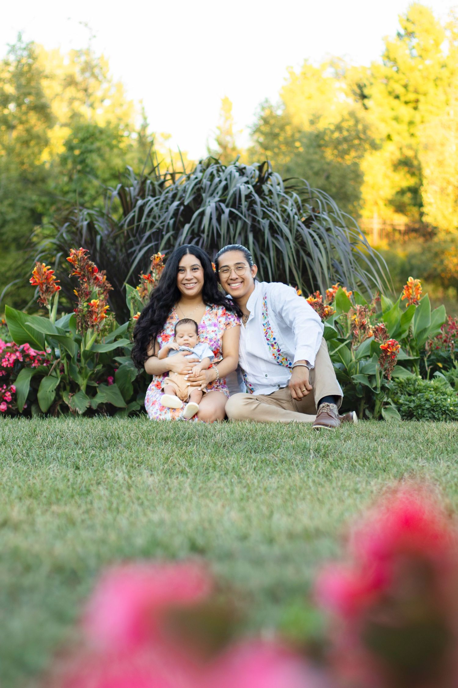 Family sits together on grass in garden with ornamental plants and flowers in the background.