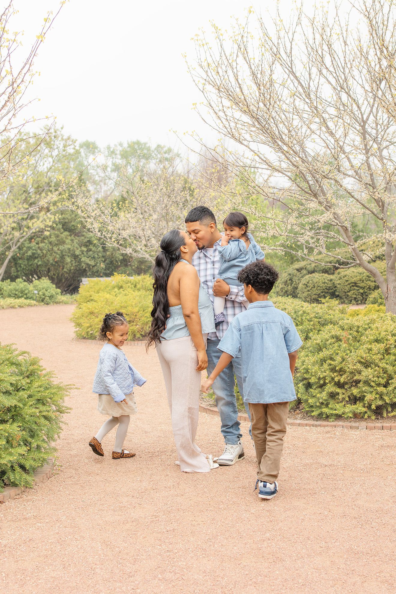 Family of five in coordinating blue and neutral outfits walking together on a garden path surrounded by green shrubs.