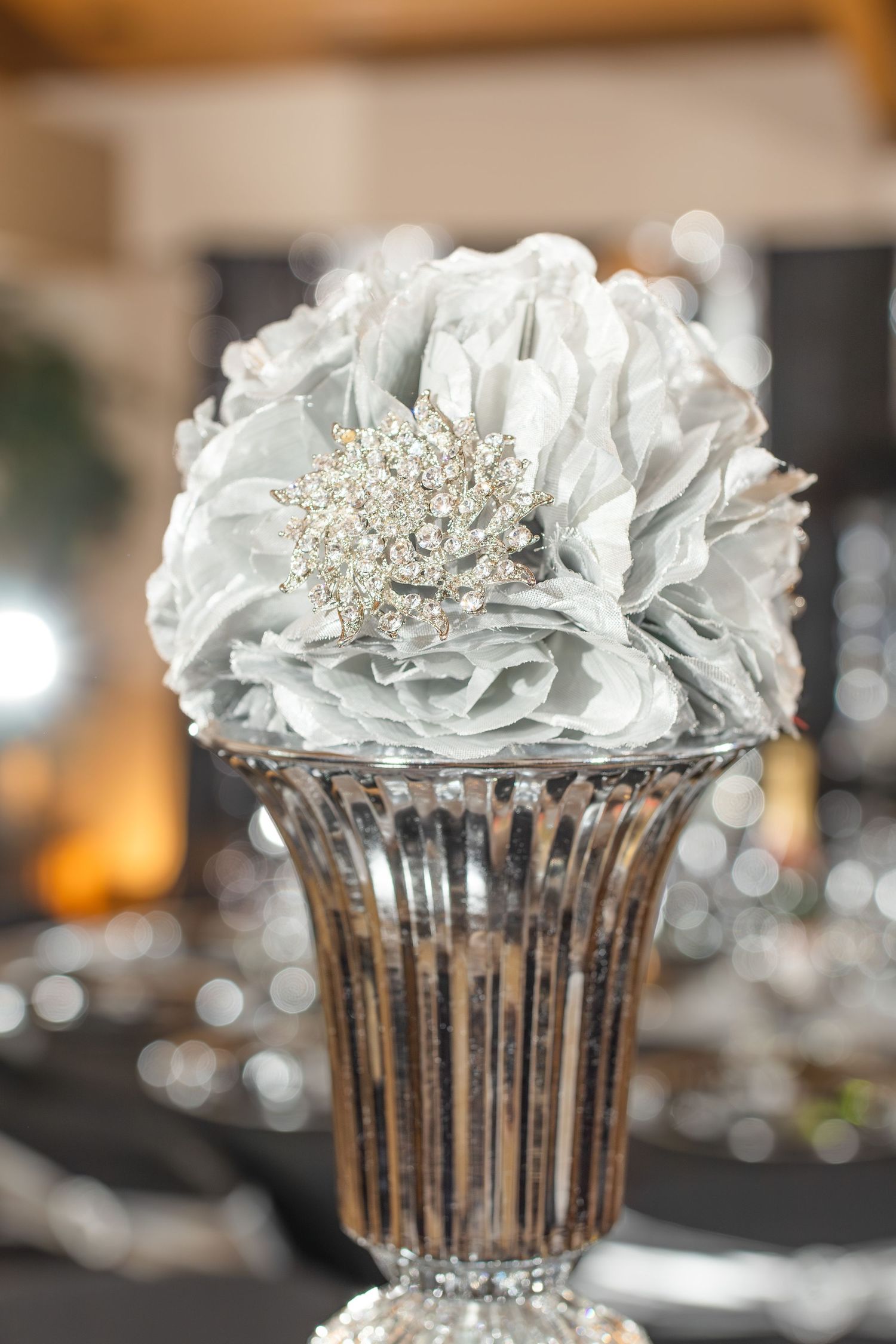 Silver fabric flower with crystal brooch centerpiece displayed in a fluted glass vase at an elegant event.