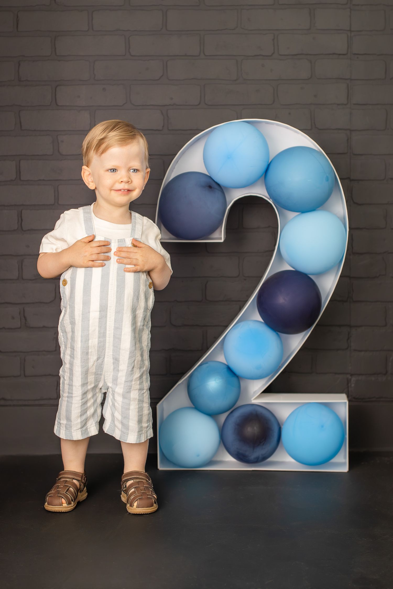 A toddler boy in striped overalls stands next to a large number 2 filled with blue balloons for his 2nd birthday.