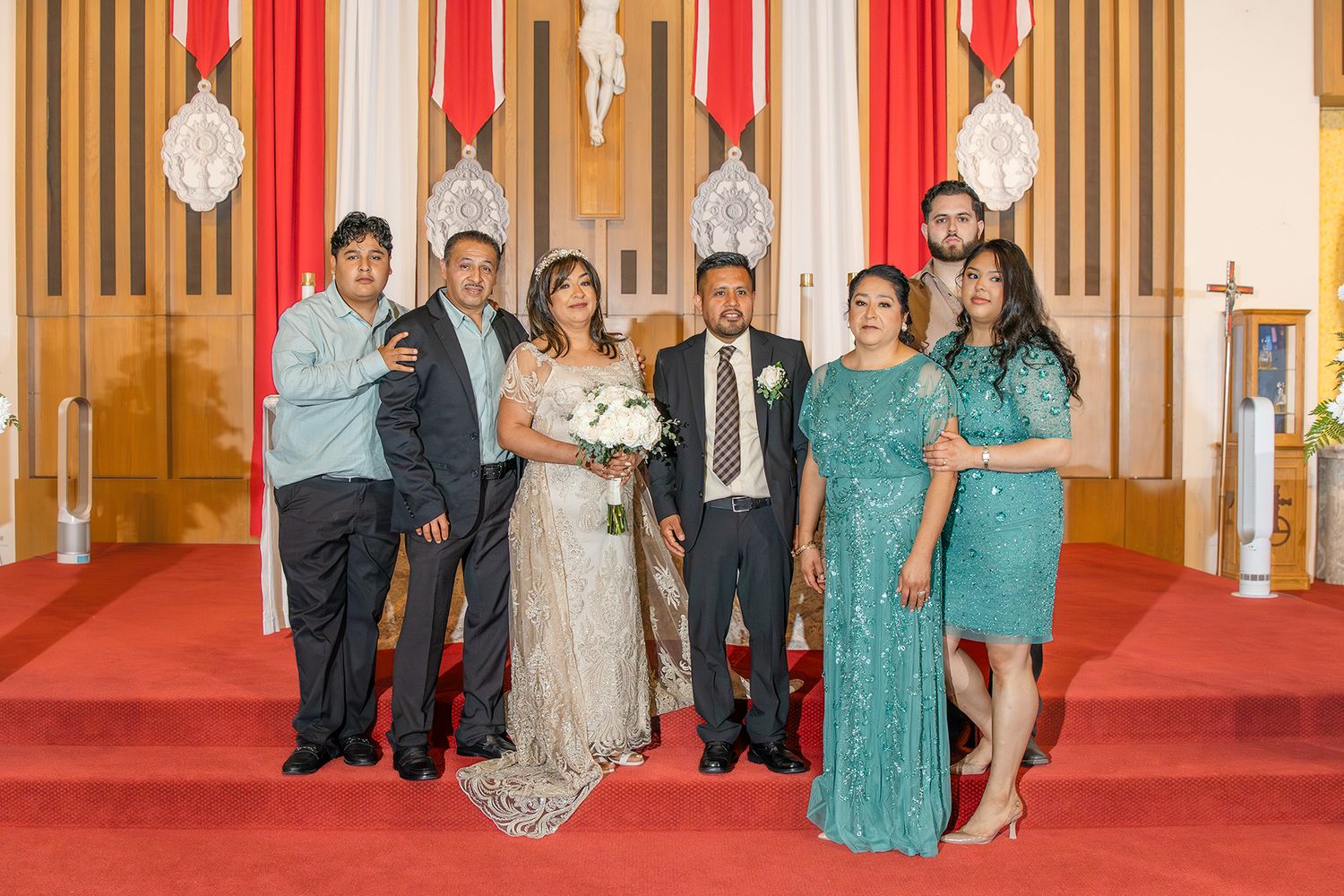 Wedding party poses in a church with red carpet, bride in lace gown holding bouquet, groom in suit, family in formal attire.