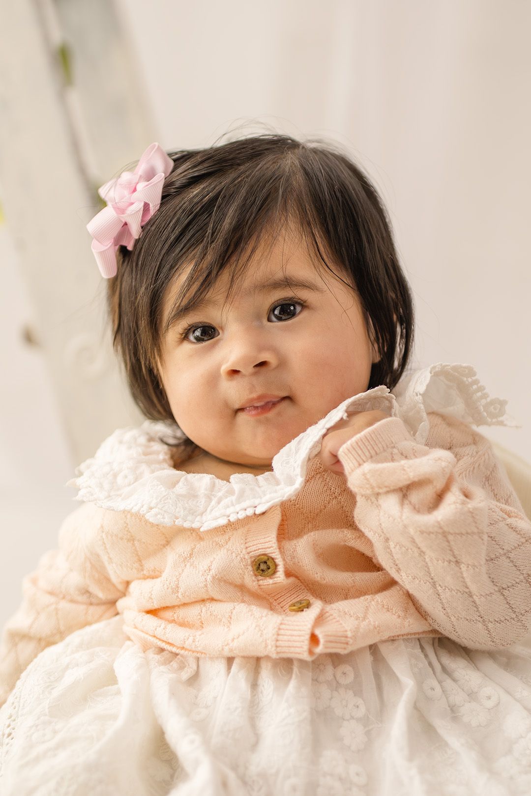 Adorable baby girl with dark hair and pink bow wearing a peach knit cardigan over a white lace dress, smiling sweetly.