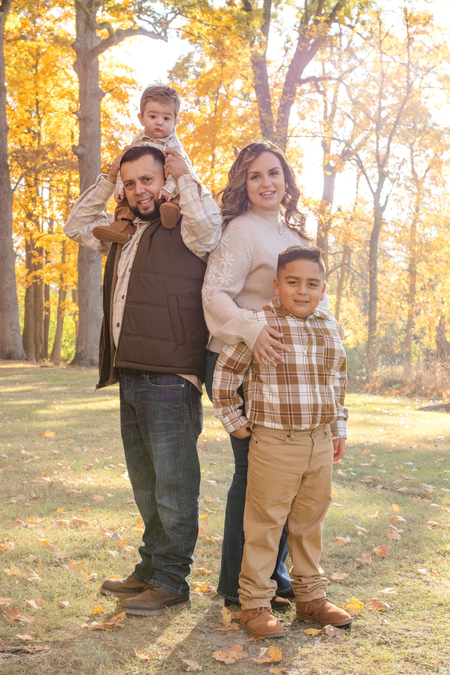 A family poses together on fallen autumn leaves with golden trees creating a warm backdrop in a park setting.