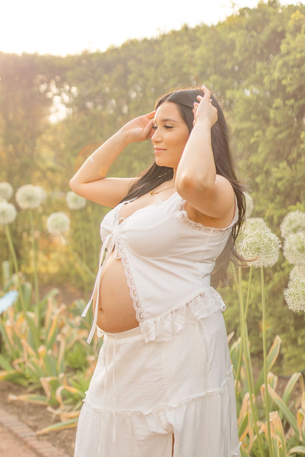 Pregnant woman in white dress poses outdoors among allium flowers during a glowing golden hour maternity shoot.