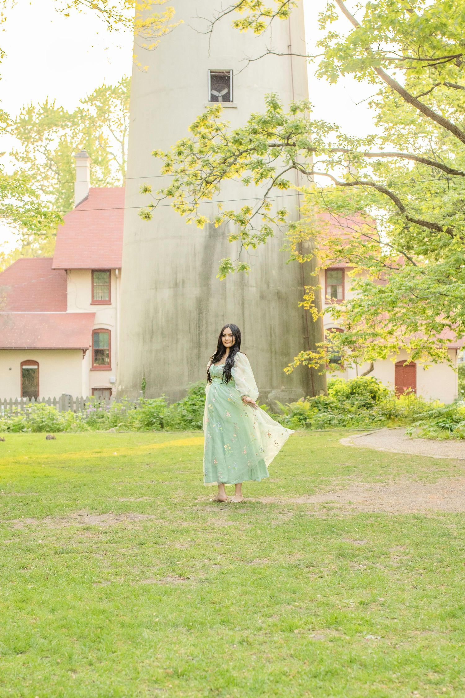 A person in a flowy mint green dress poses gracefully on grass near a lighthouse with red-roofed buildings.