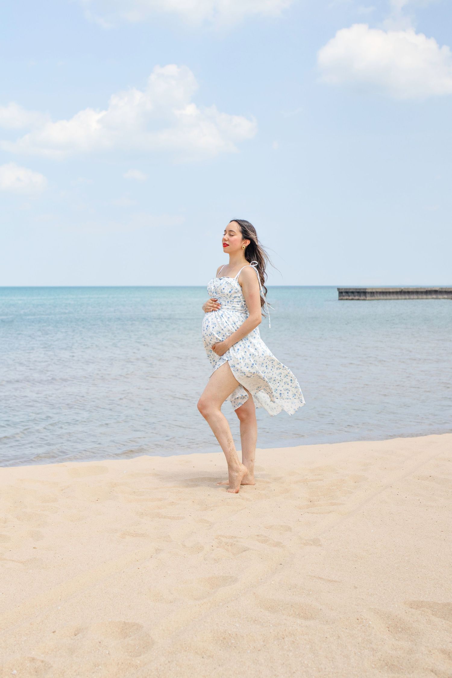 Woman in white dress joyfully twirls on a serene beach with turquoise waters and sandy shoreline visible.