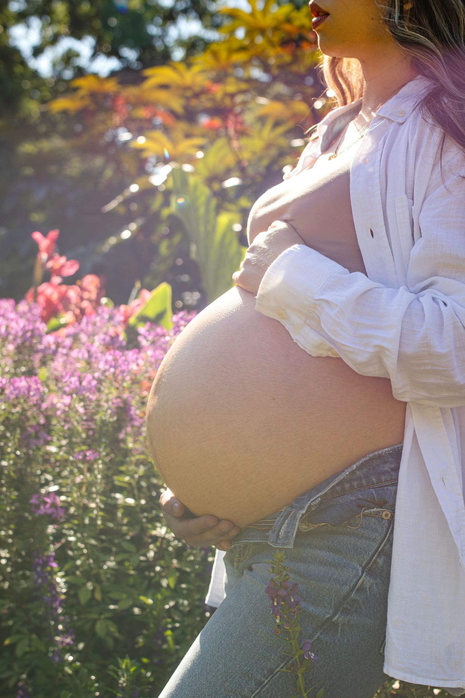 A silhouette of a pregnant belly against a sunlit garden background with pink flowers and autumn foliage.