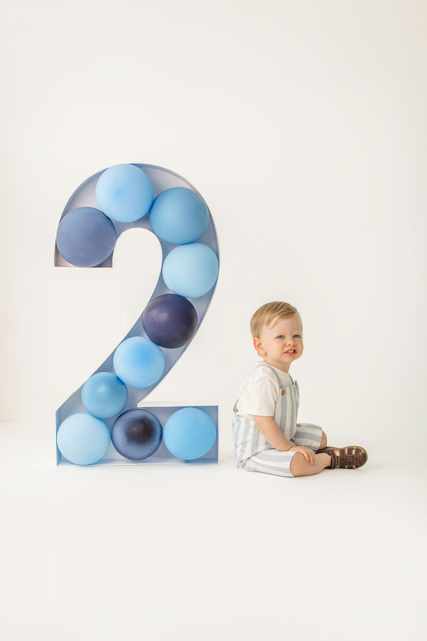 Toddler boy sitting beside a large number 2 decorated with blue balloons on white background for 2nd birthday.