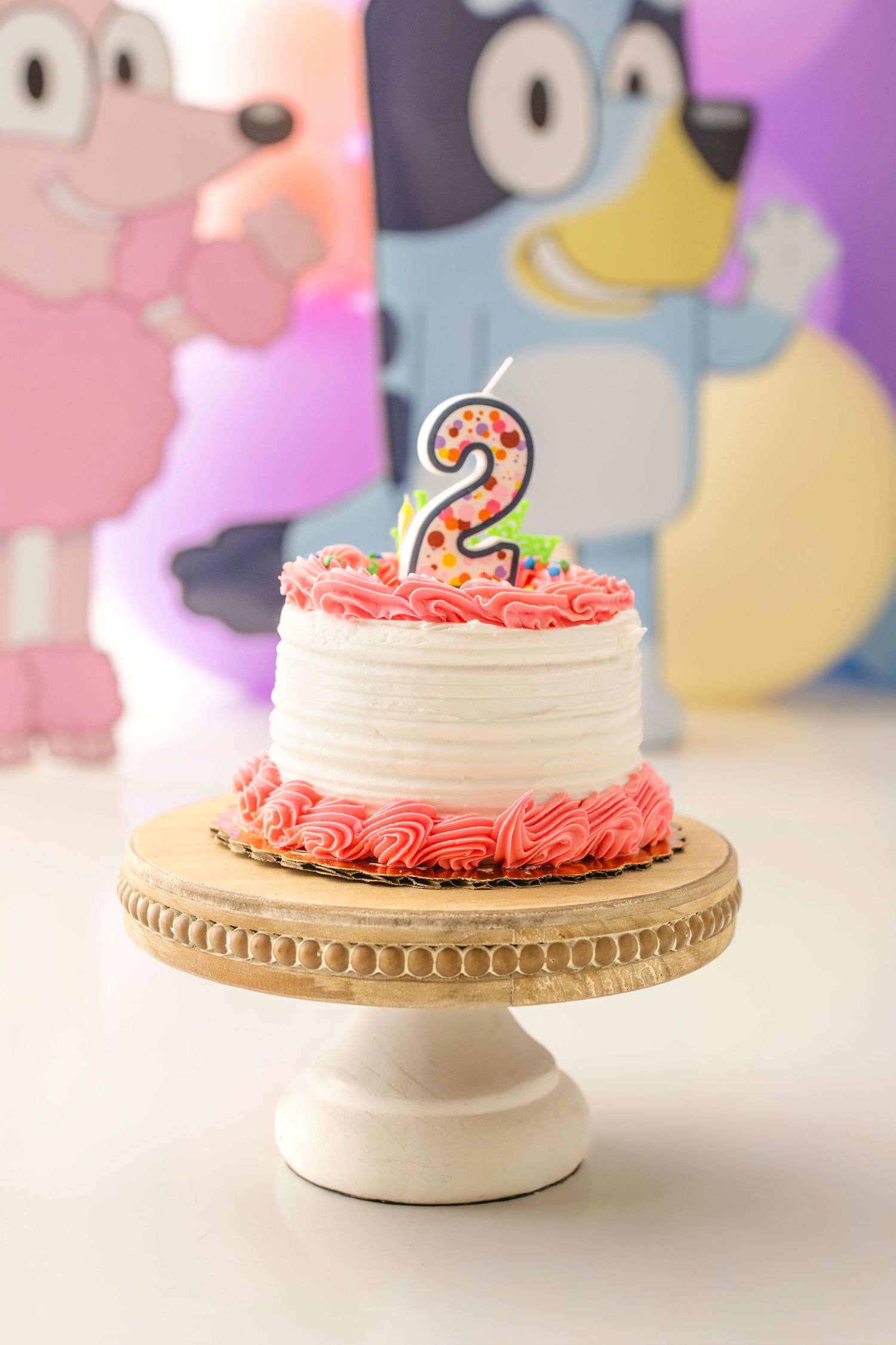 A white and pink 2nd birthday cake with a number 2 candle on a wooden stand, with Bluey cartoon backdrop.