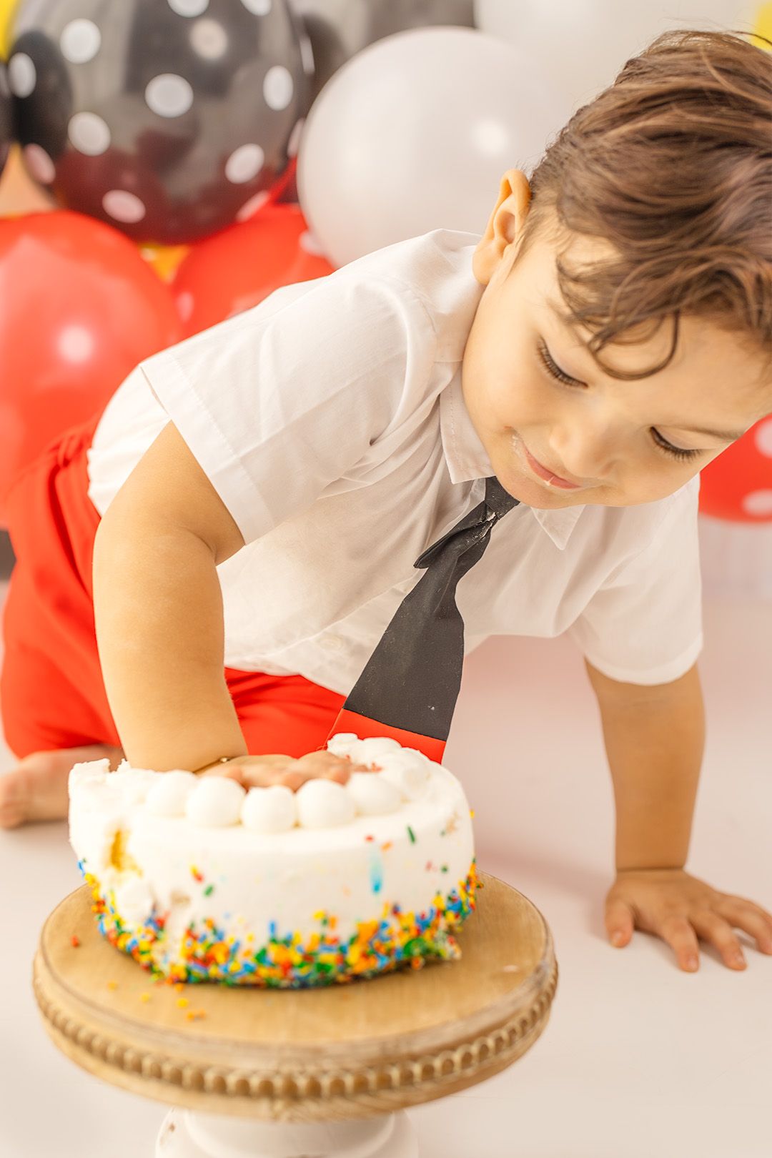 Young boy in white shirt and black tie leaning over colorful sprinkle birthday cake at party.