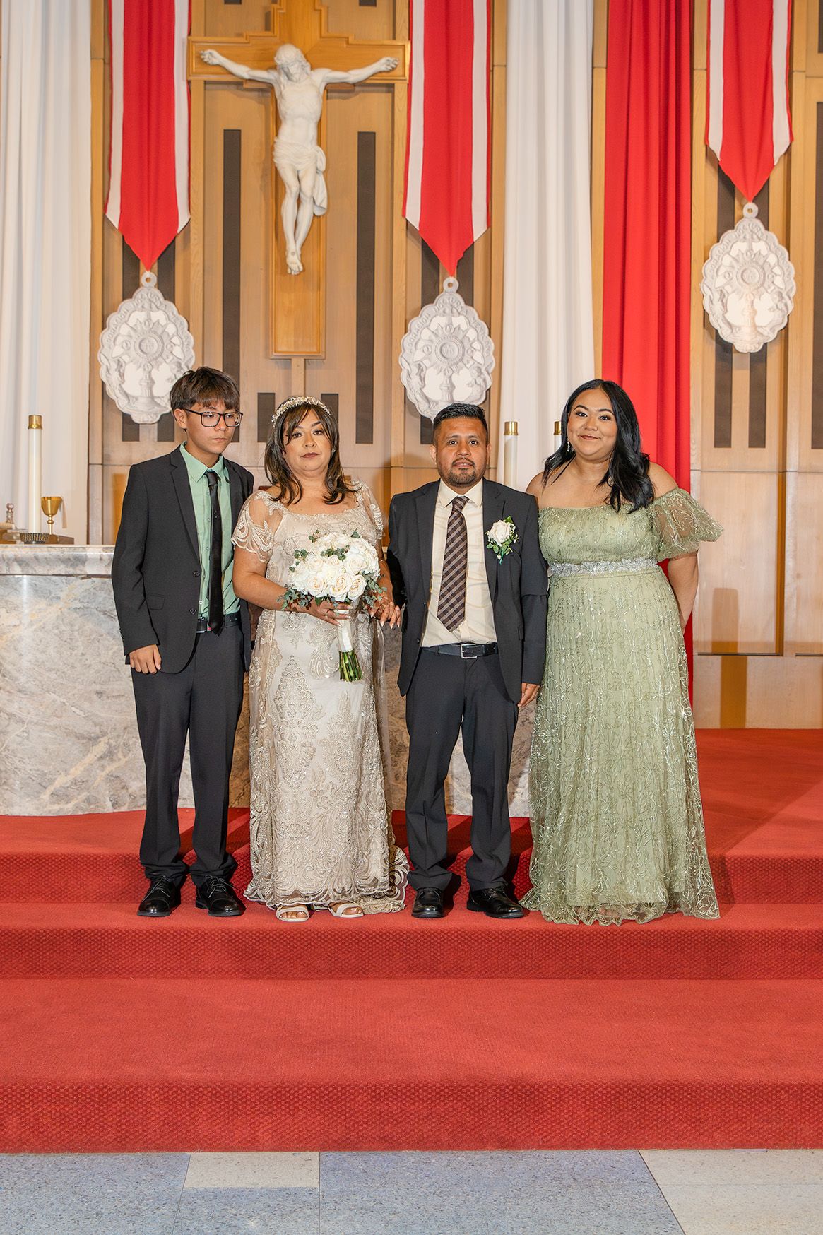 Family poses at church altar during wedding ceremony, bride in lace gown holding white bouquet with cross backdrop.