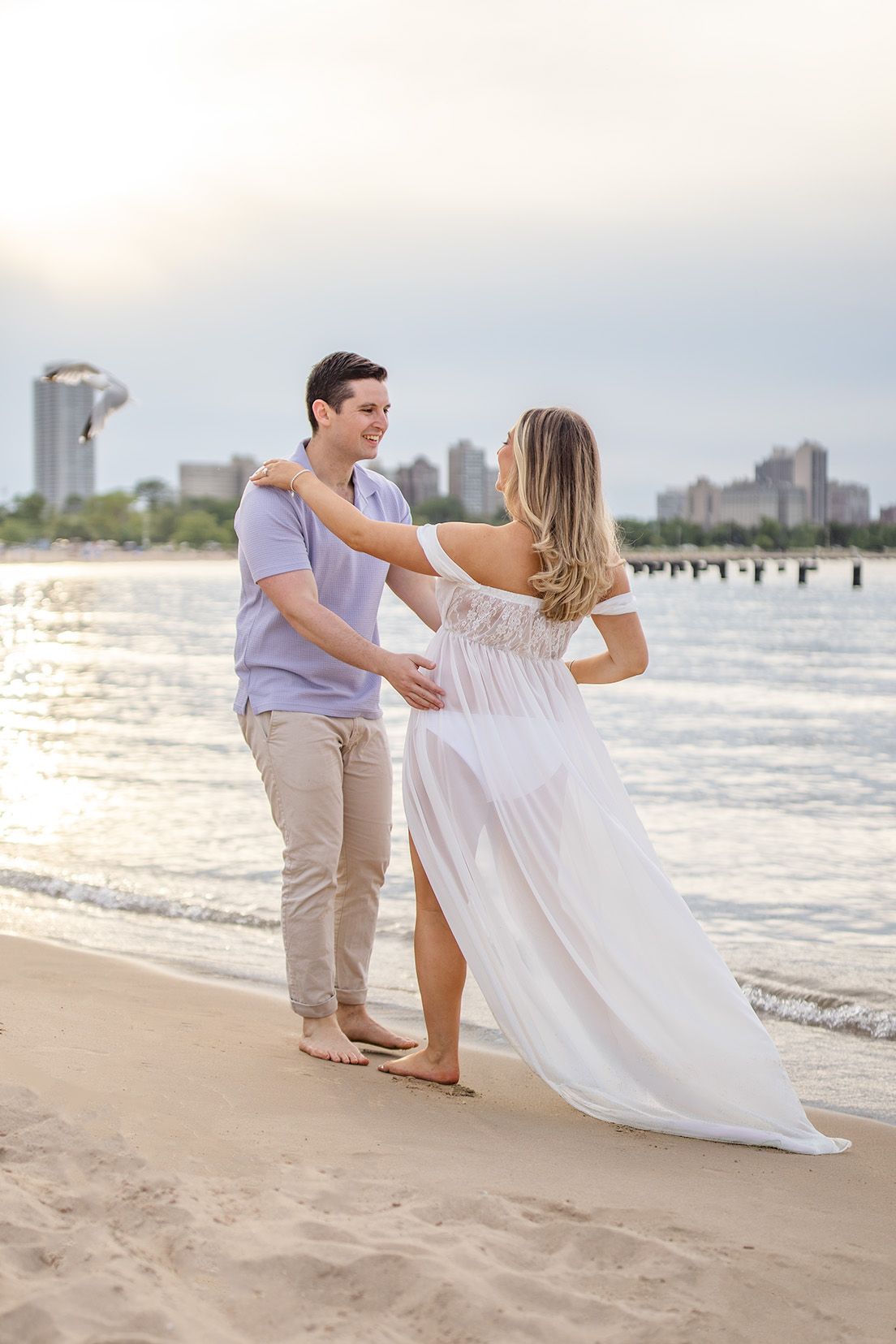 Couple embracing on sandy beach at sunset, woman in white off-shoulder gown, city skyline in background.