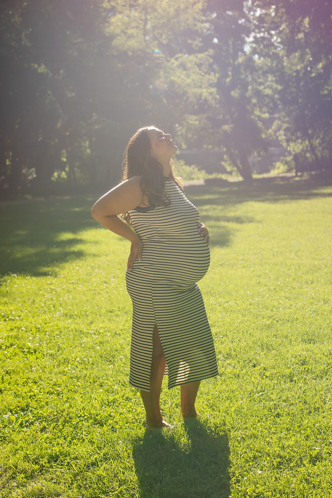 A pregnant woman in a striped maxi dress stands on green grass, glowing in warm sunlight with hands on her bump.