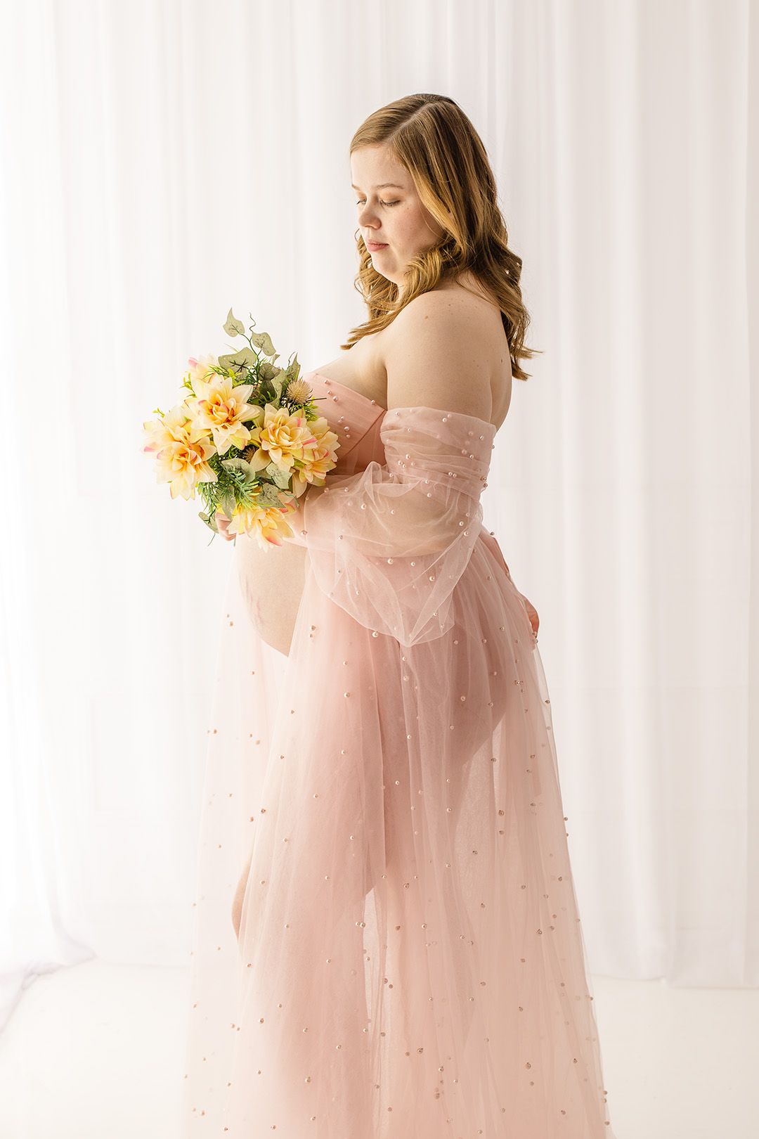 Elegant maternity portraits featuring flowing blush pink gown with pearl details and yellow floral bouquet.