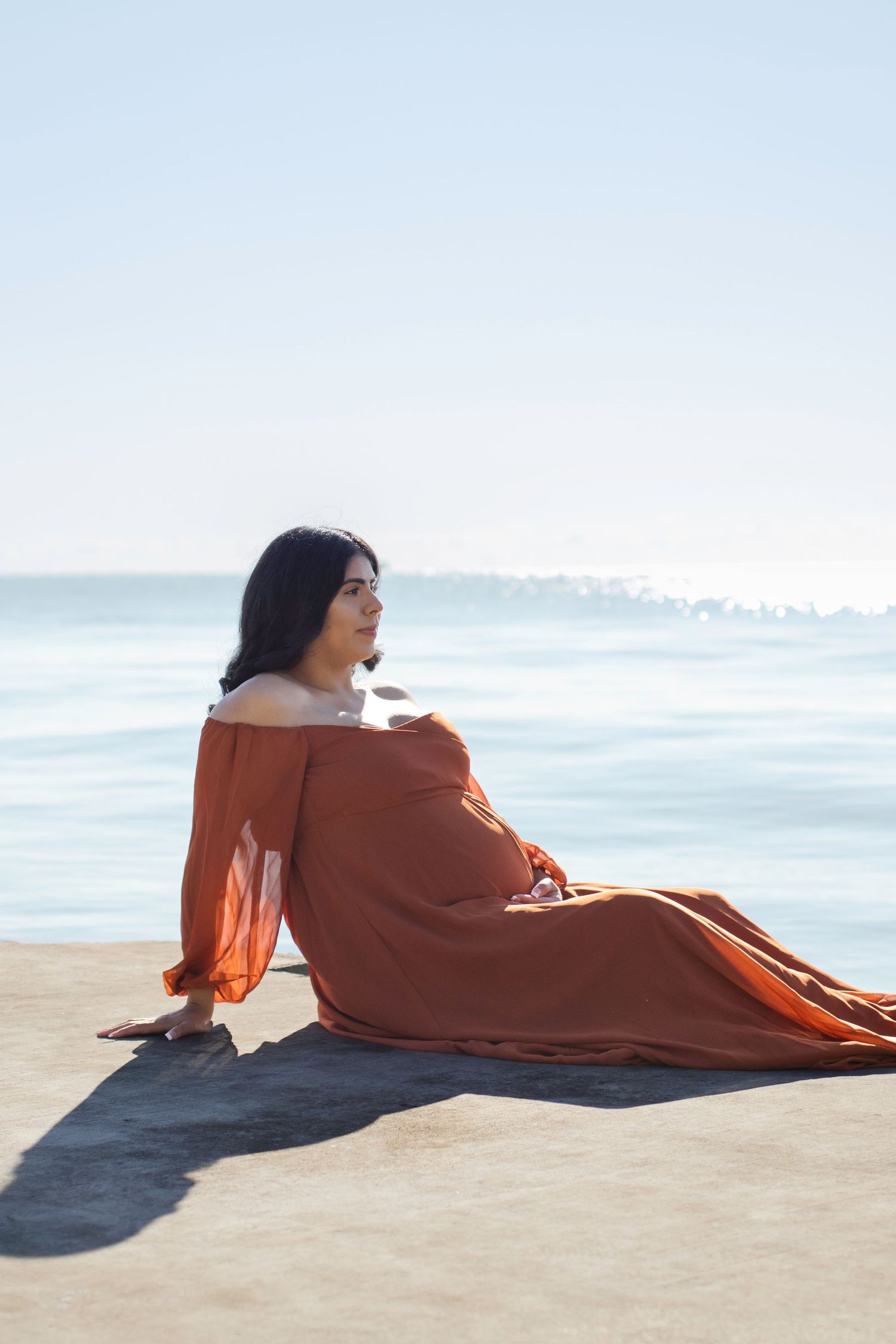 Maternity photo of person in rust-colored dress sitting on beach at sunset.