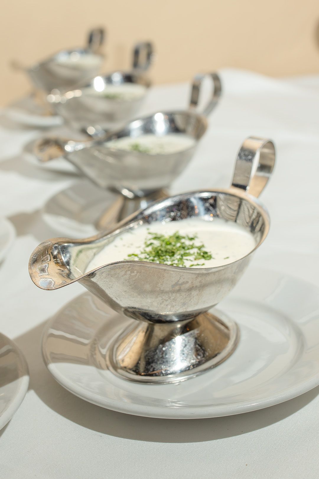 Silver gravy boats filled with creamy white sauce garnished with fresh chives on white plates.