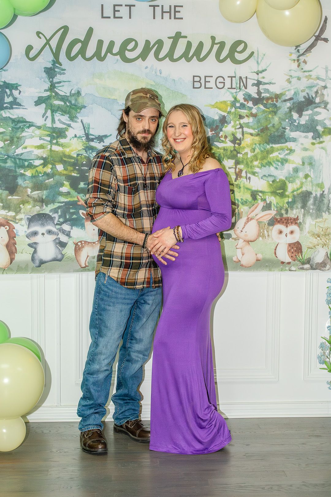 A couple poses at a woodland-themed baby shower with 'Let the Adventure Begin' backdrop and green and yellow balloons.