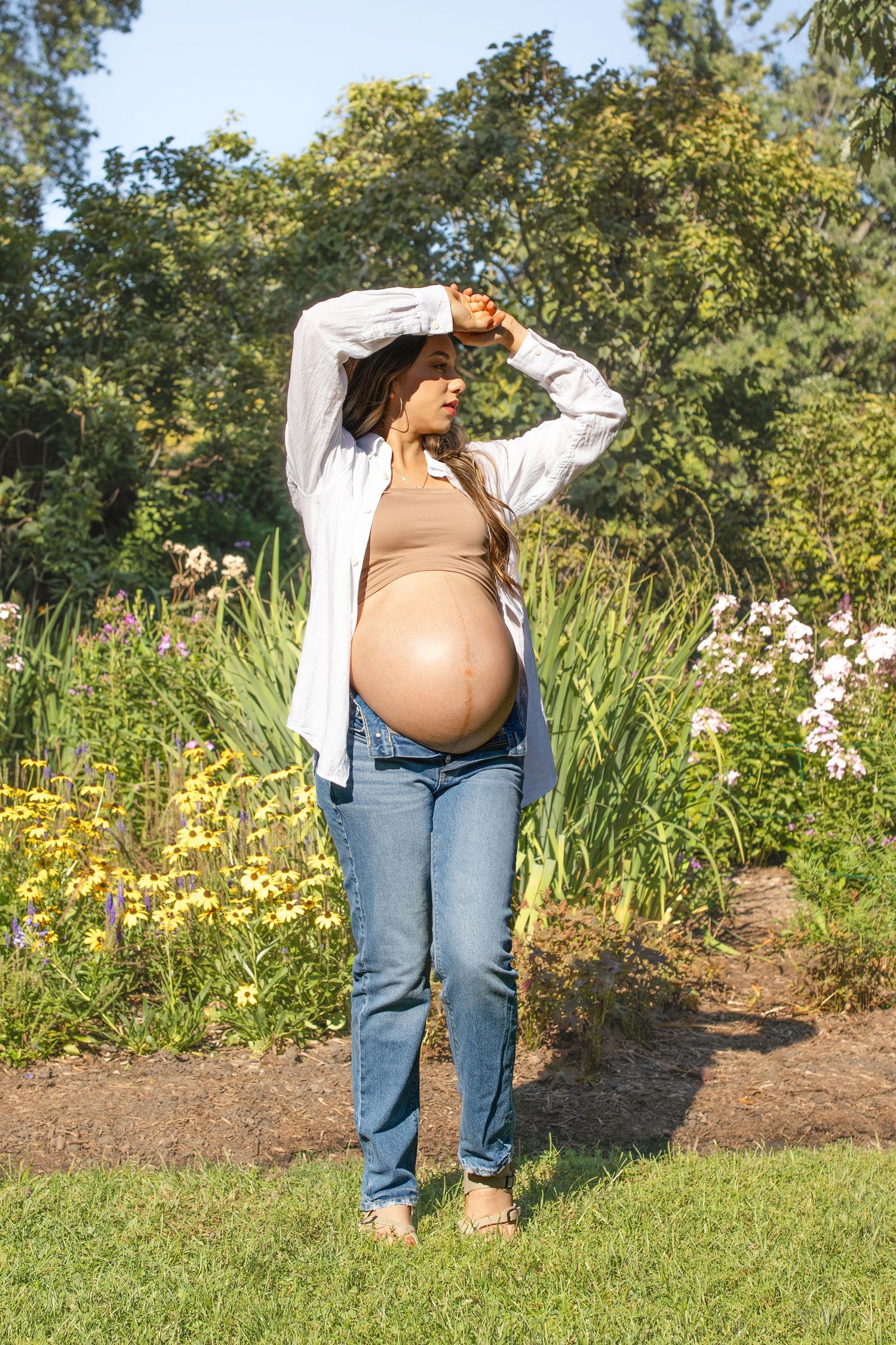 A pregnant person in jeans and an open white shirt poses in front of a garden filled with colorful flowers and greenery.