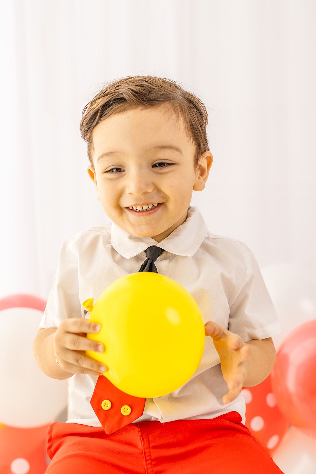 Smiling toddler boy holding a yellow balloon, wearing a white shirt with black tie and red pants at a birthday party.