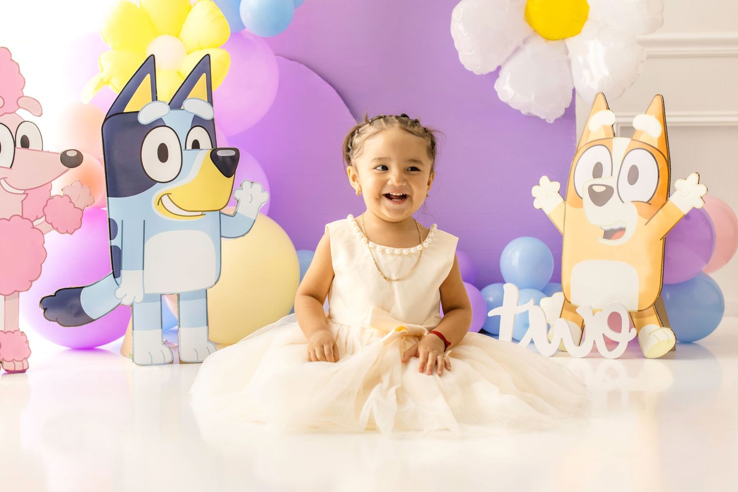 Smiling toddler girl in white dress at Bluey themed first birthday party with colorful balloon backdrop.