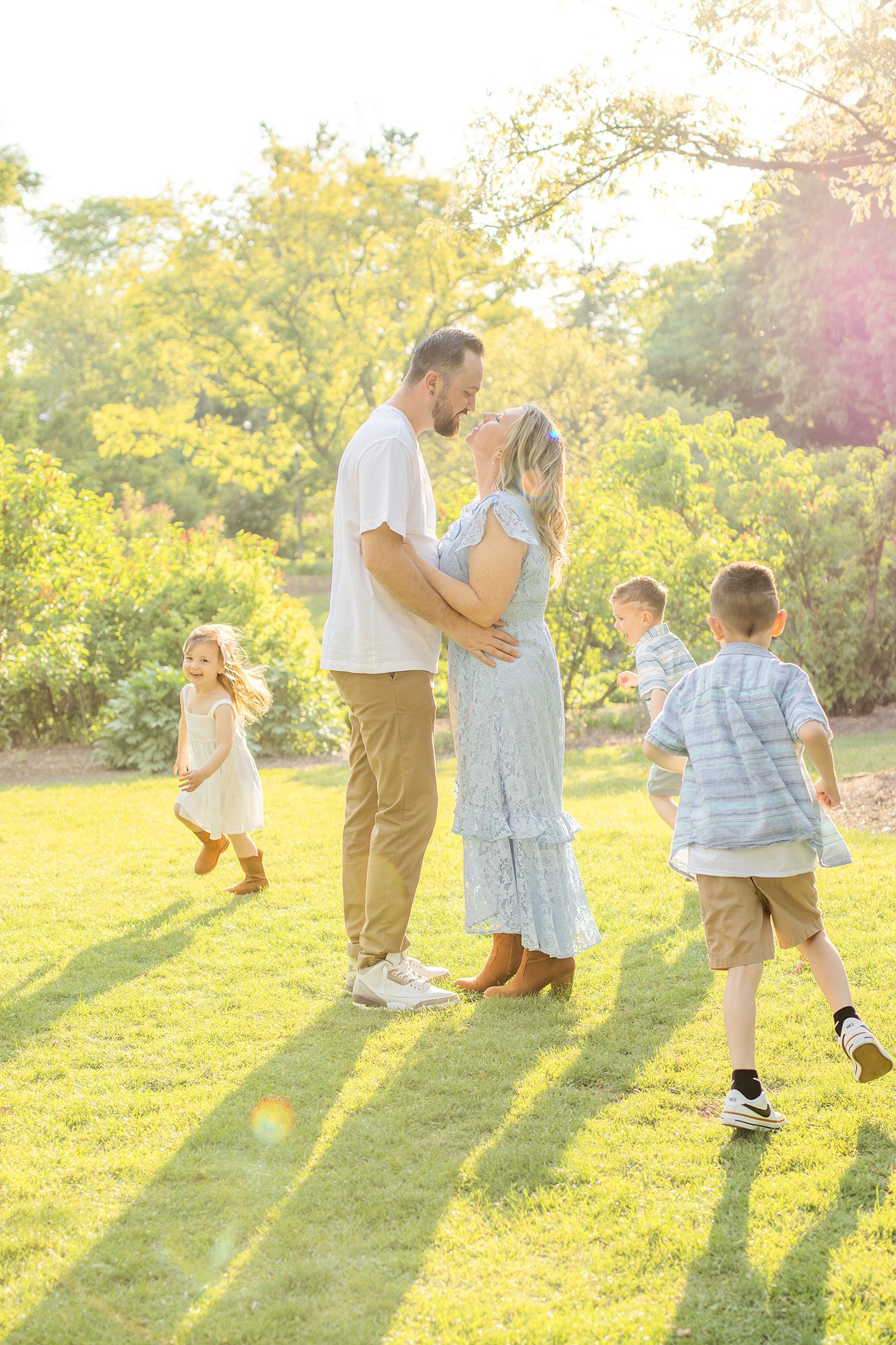 A couple shares a kiss while their three children play around them in a sunlit green garden.