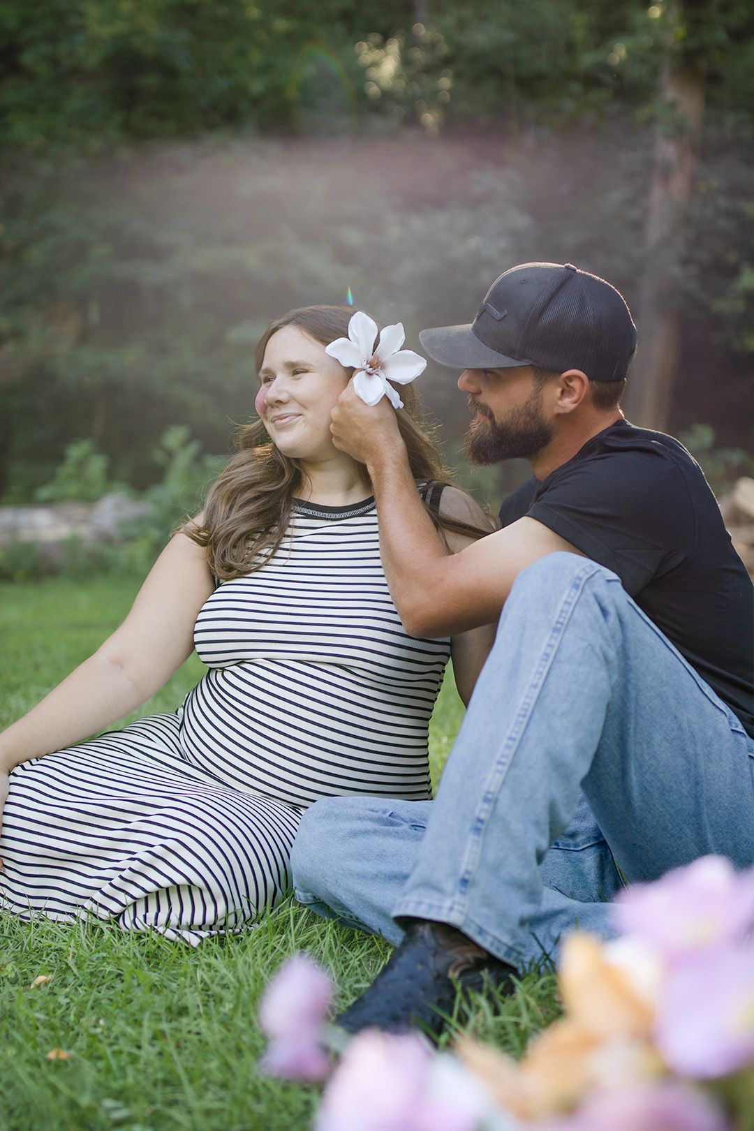 A smiling couple sits on grass as a man places a flower in a pregnant woman's hair outdoors.