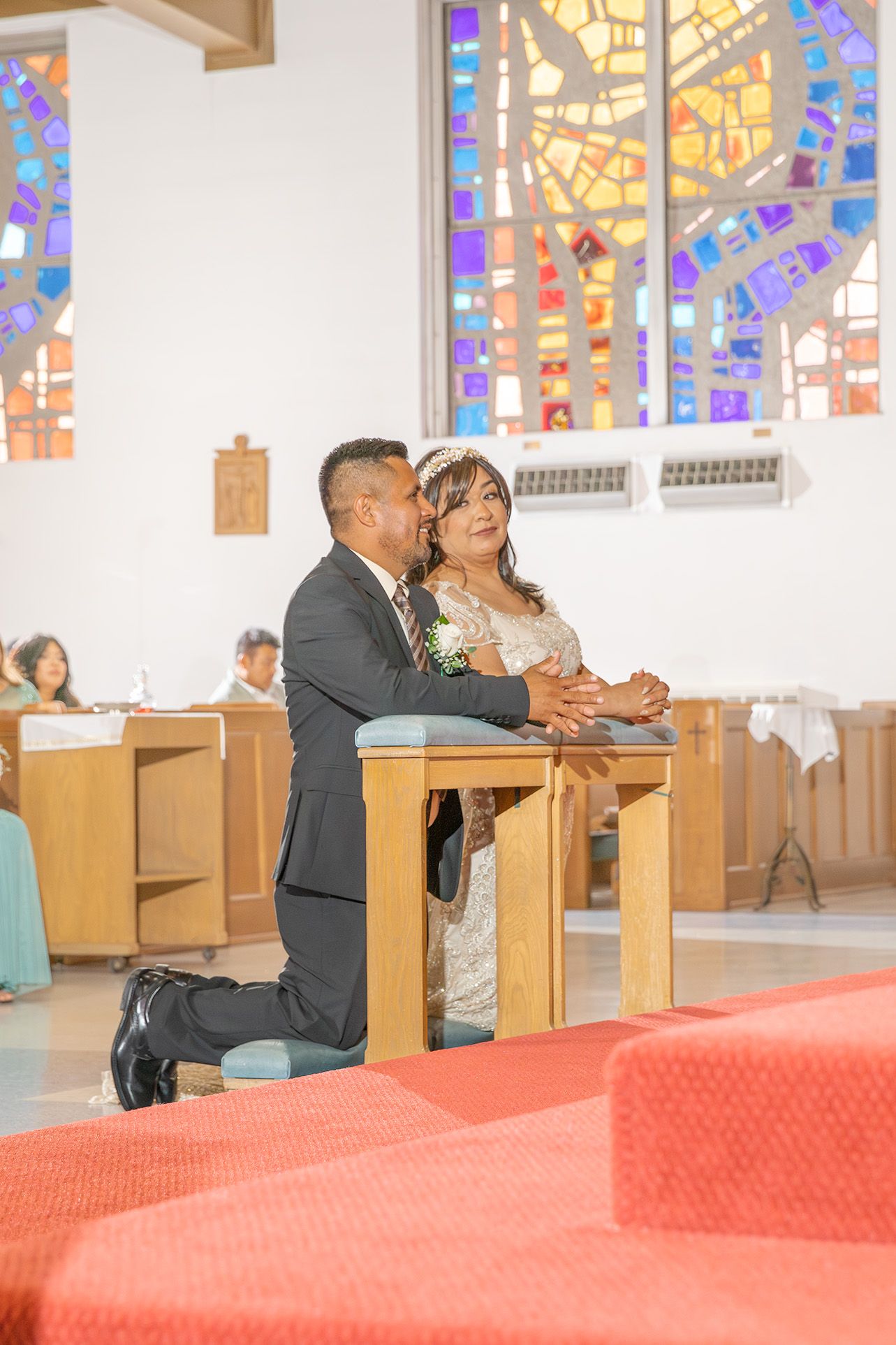 Bride and groom kneeling at church altar during wedding ceremony with colorful stained glass windows behind them.