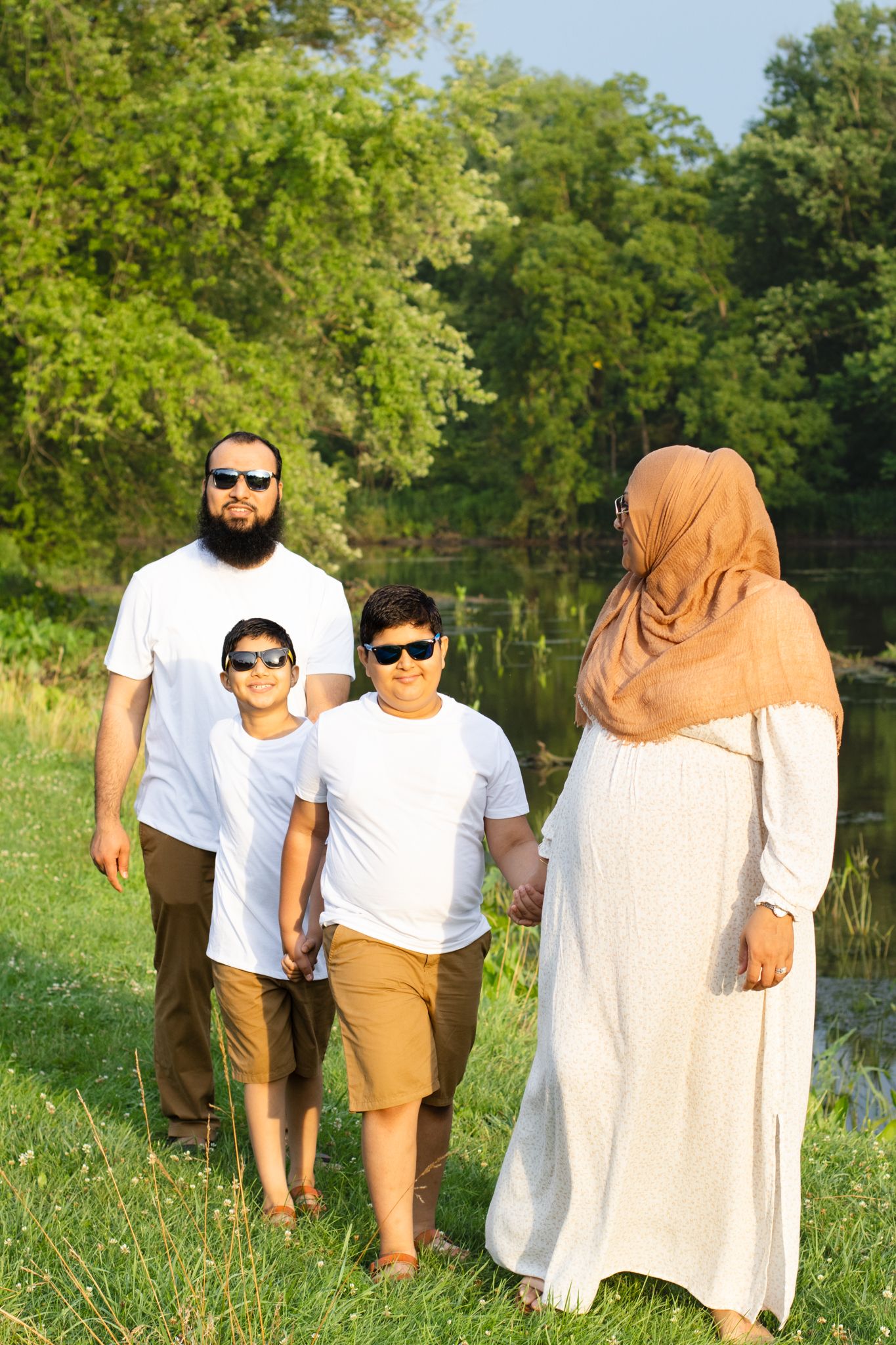 Family walks together along grassy path near pond in wooded park.