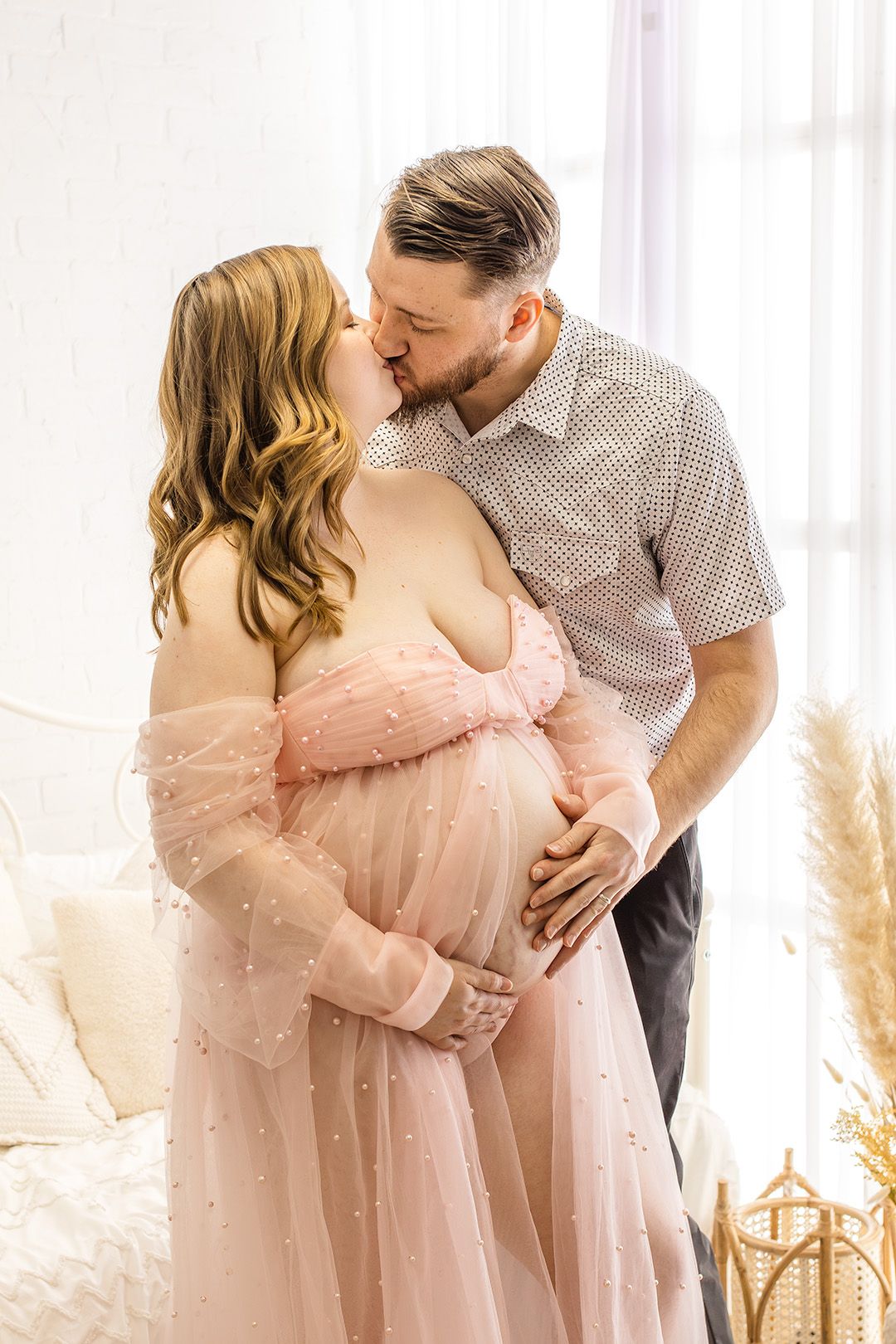 Intimate maternity moment captured with couple in pink gown and button-down shirt against white curtain backdrop.