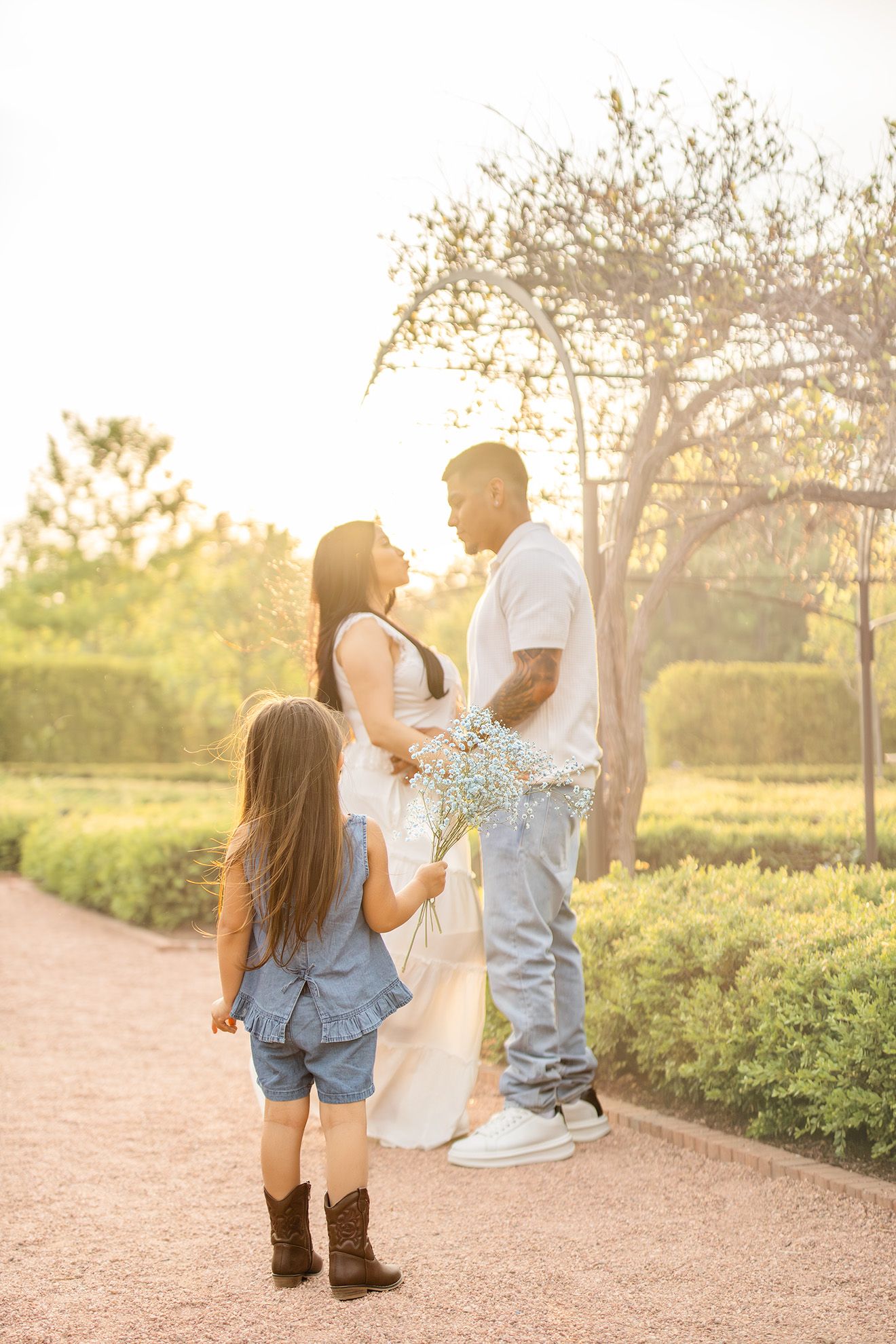 Family maternity photo with pregnant woman, man, and young girl with flowers in sunlit garden path.