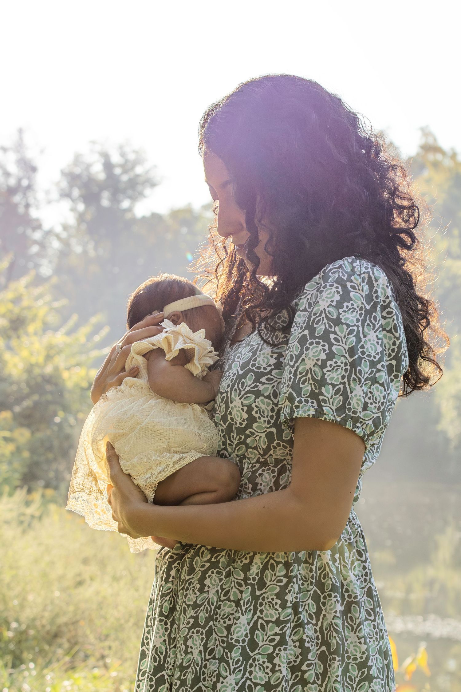 Mother in floral dress cradles sleeping newborn against her chest in golden afternoon light.