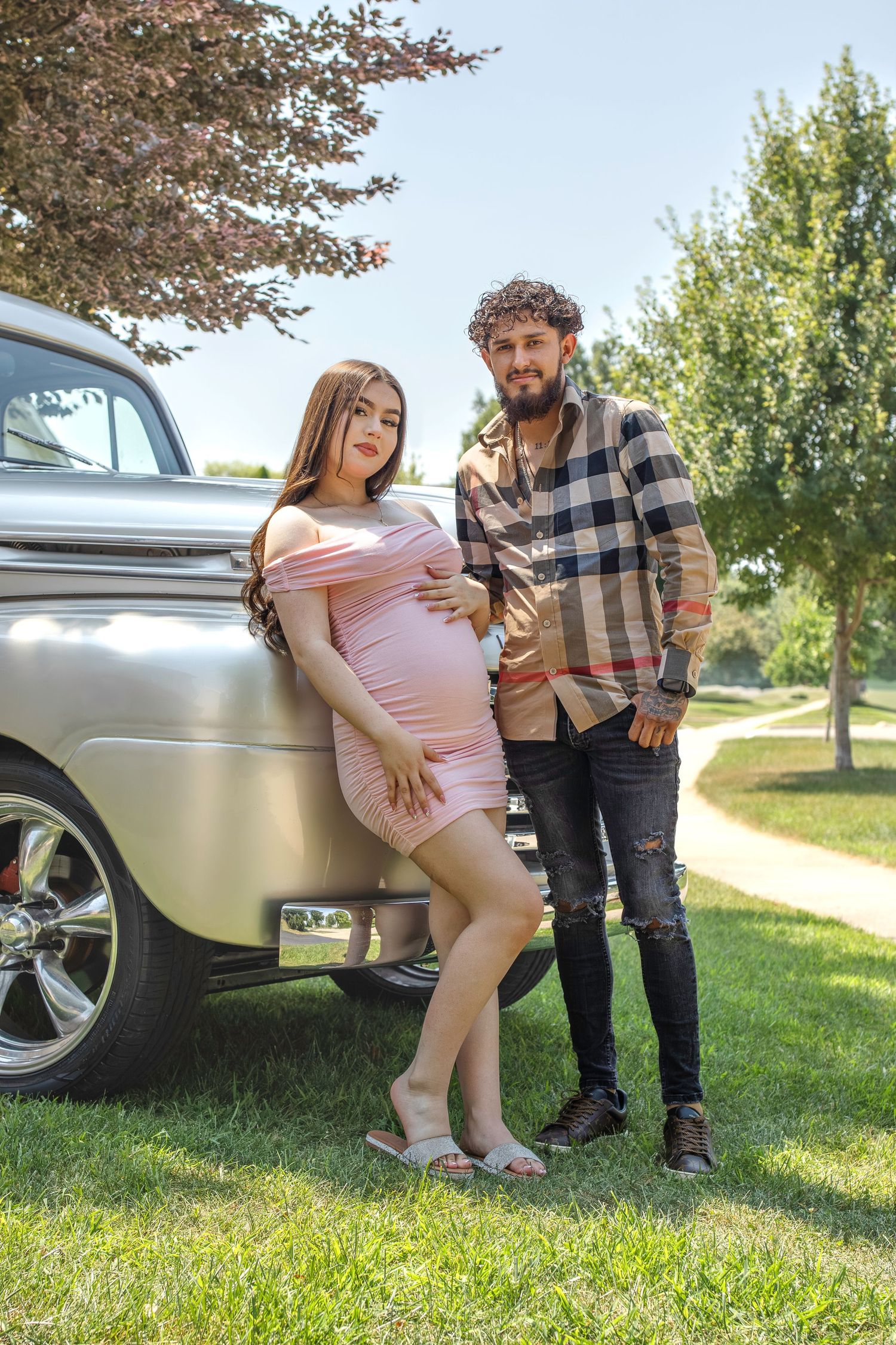 A pregnant woman in pink dress leans against a vintage silver truck while standing with companion in plaid shirt outdoors.