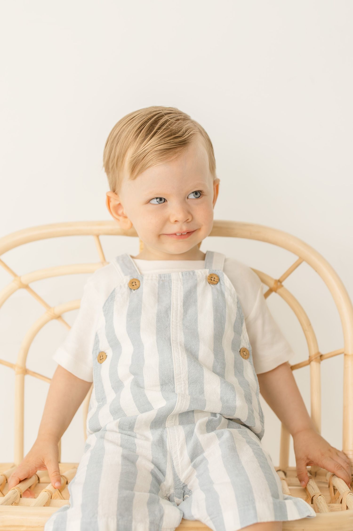 Smiling toddler boy in blue striped linen overalls and white tee sits on a rattan chair.